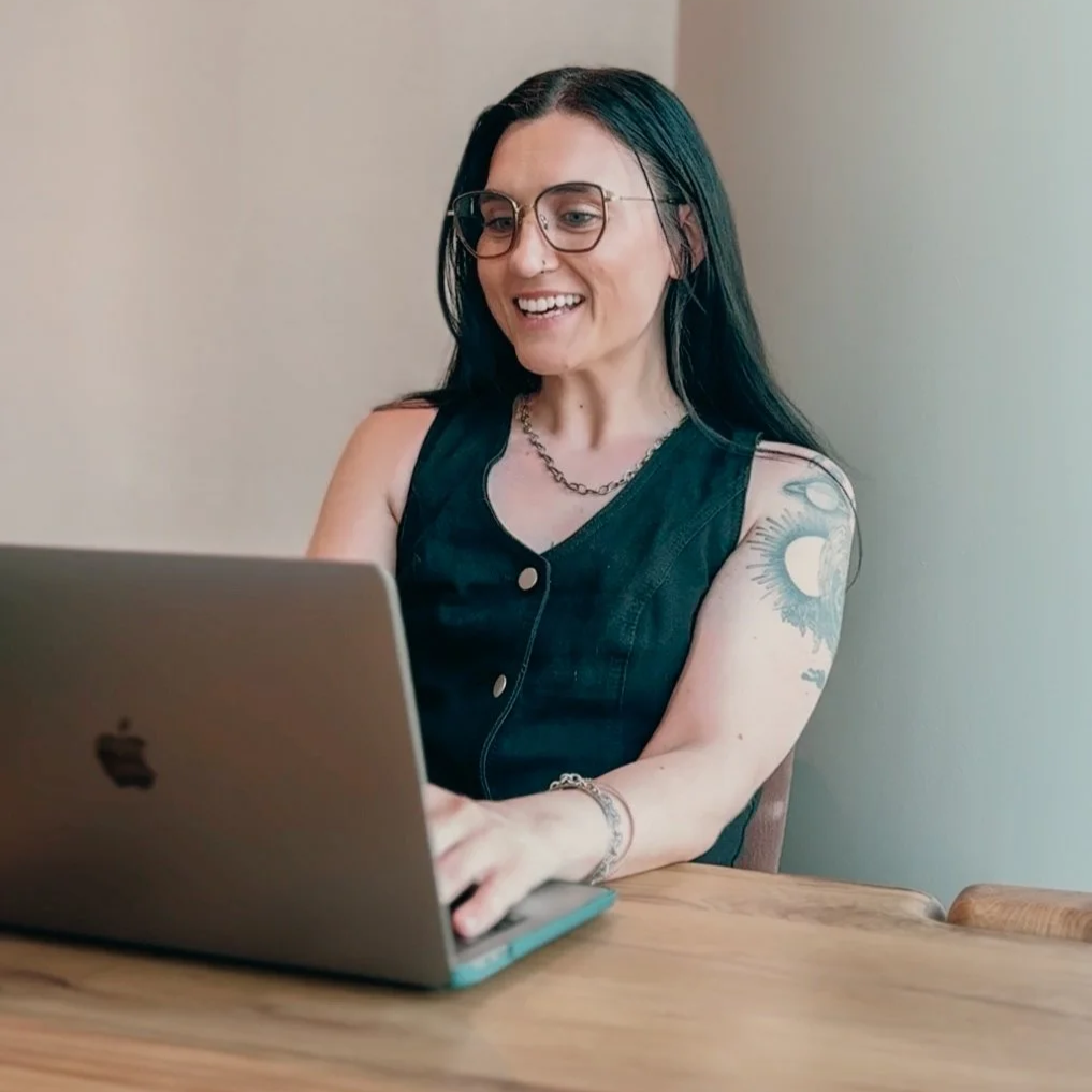 A woman with black hair, glasses, and tattoos on her arm, sitting at a wooden table and working on a silver MacBook laptop, smiling and looking at the screen.