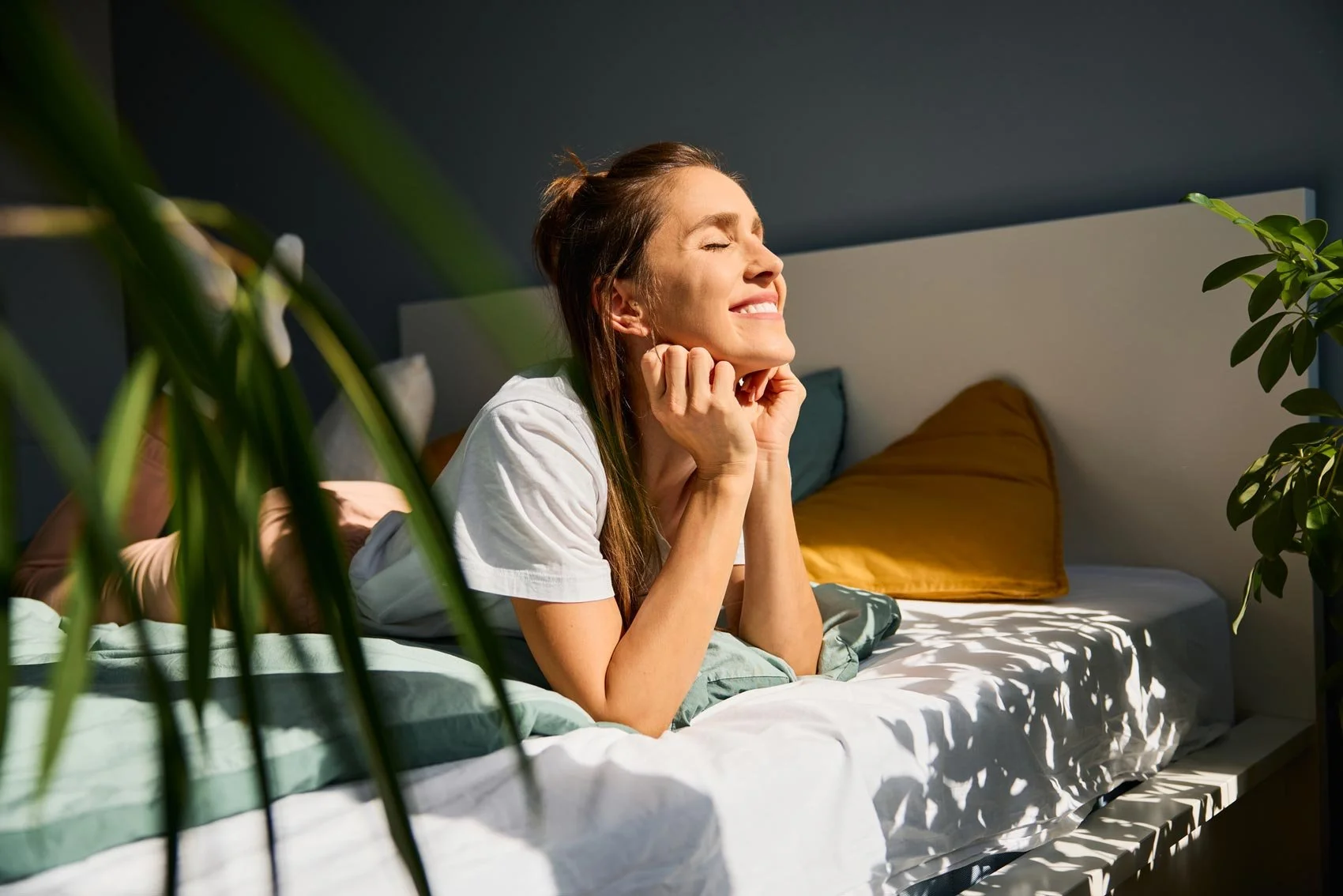 Woman lying on bed with closed eyes, smiling, in a well-lit bedroom with plants and colorful pillows.