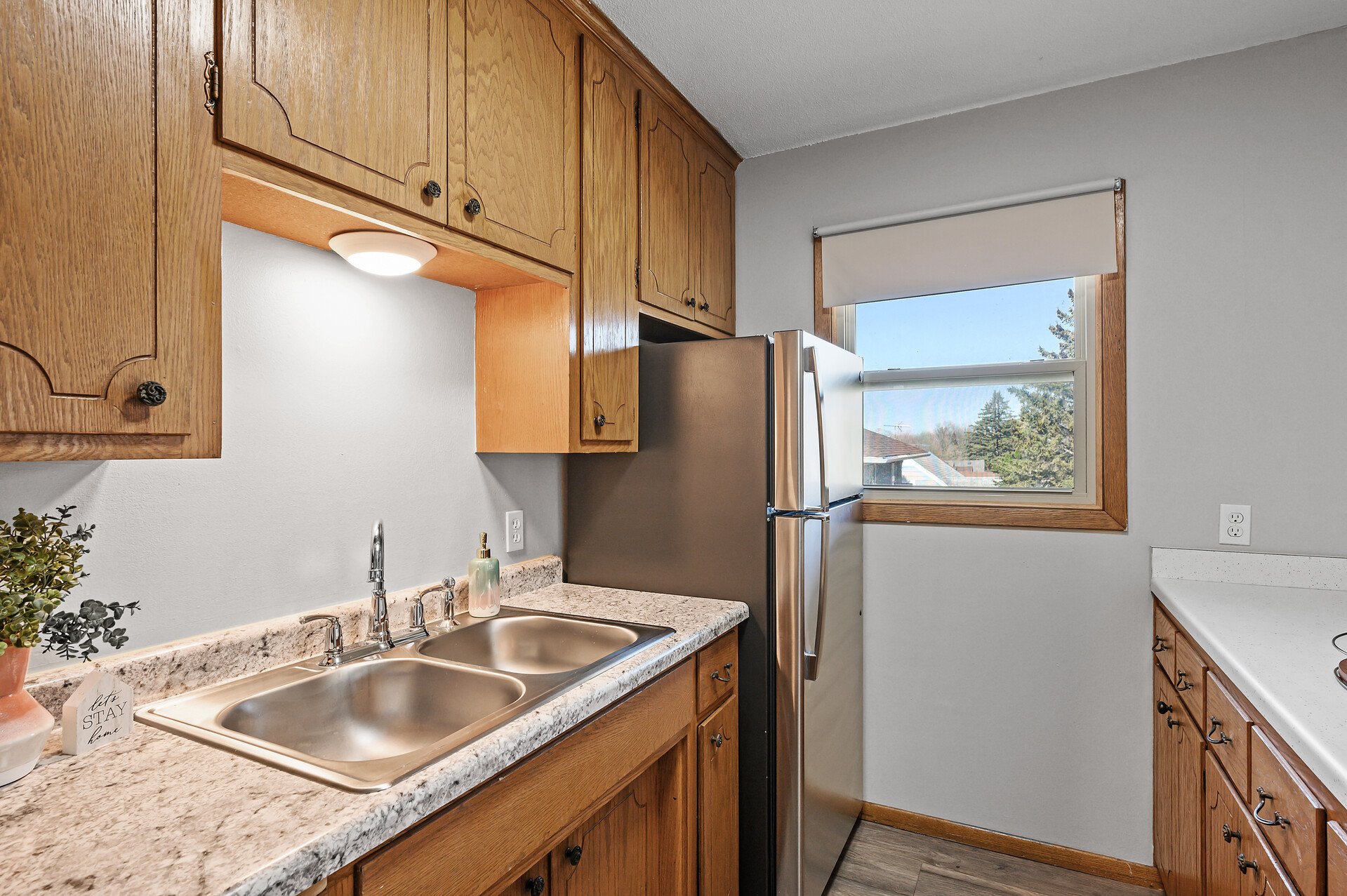 A kitchen with wooden cabinets, a beige granite countertop, a stainless steel double sink, a silver refrigerator, a window with a white roller shade, and a potted plant on the counter.