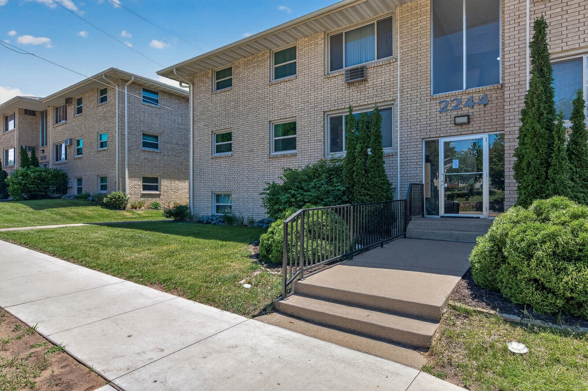 A multi-unit apartment building with a brick exterior, surrounded by green grass and bushes. The entrance has a small sidewalk with stairs and a handrail leading to a glass door. The building has multiple windows, some with an air conditioning unit, and the number 2244 displayed near the entrance. There are trees and a clear blue sky in the background.