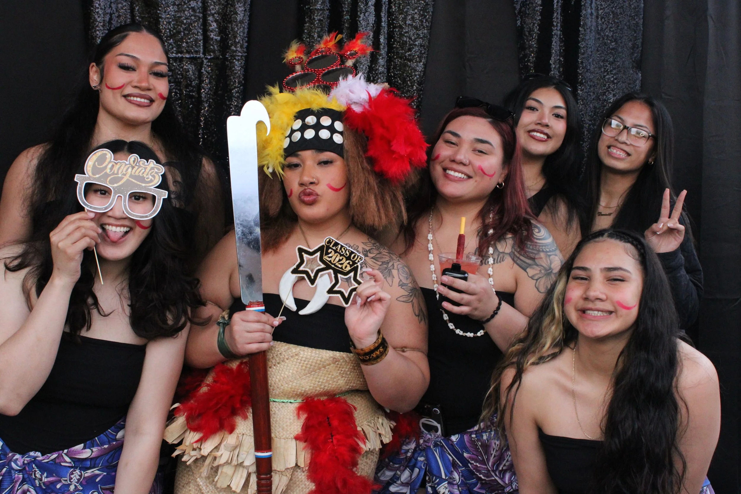 Group of young women celebrating, with some wearing costumes and face paint, holding party props, and smiling against a black, sparkly backdrop.