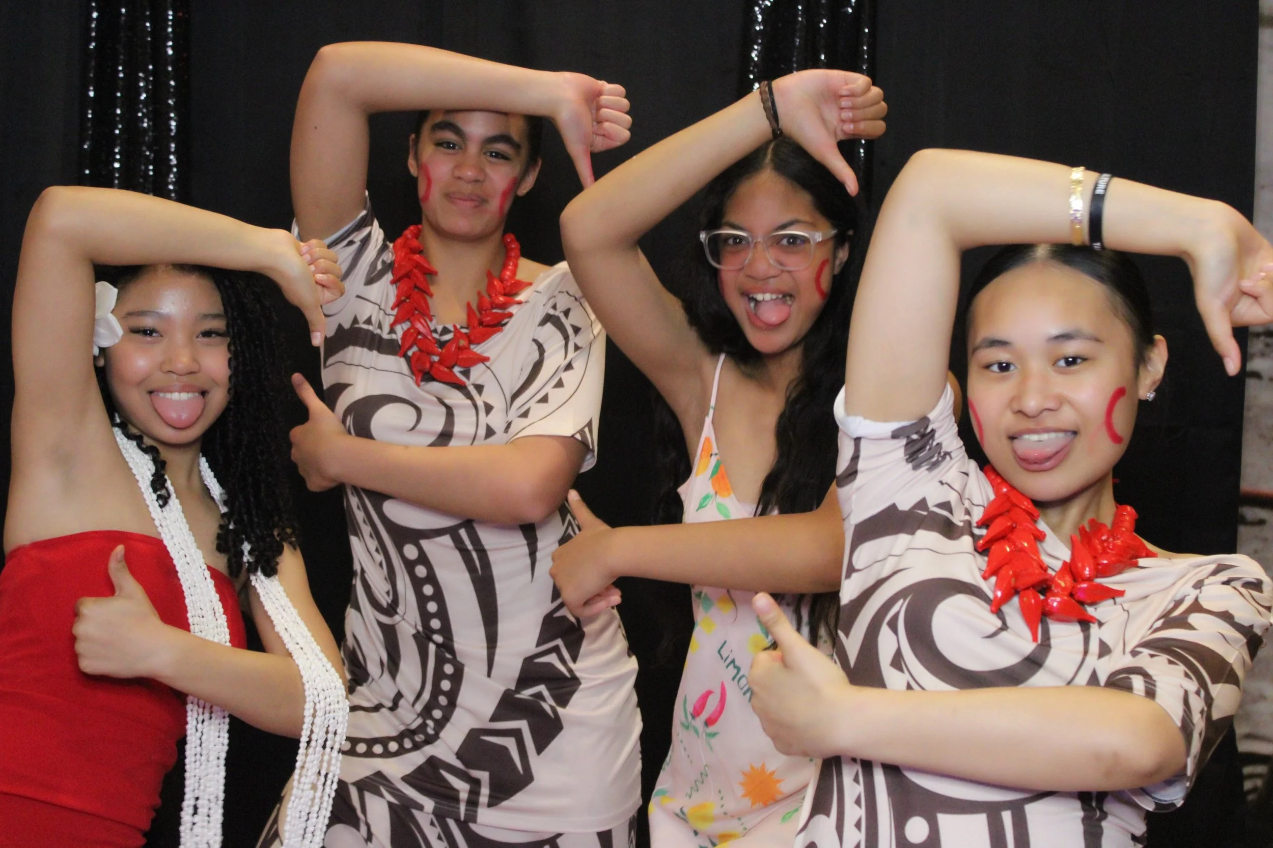 Group of four young women posing with playful expressions, wearing colorful outfits and accessories, standing against a black background.