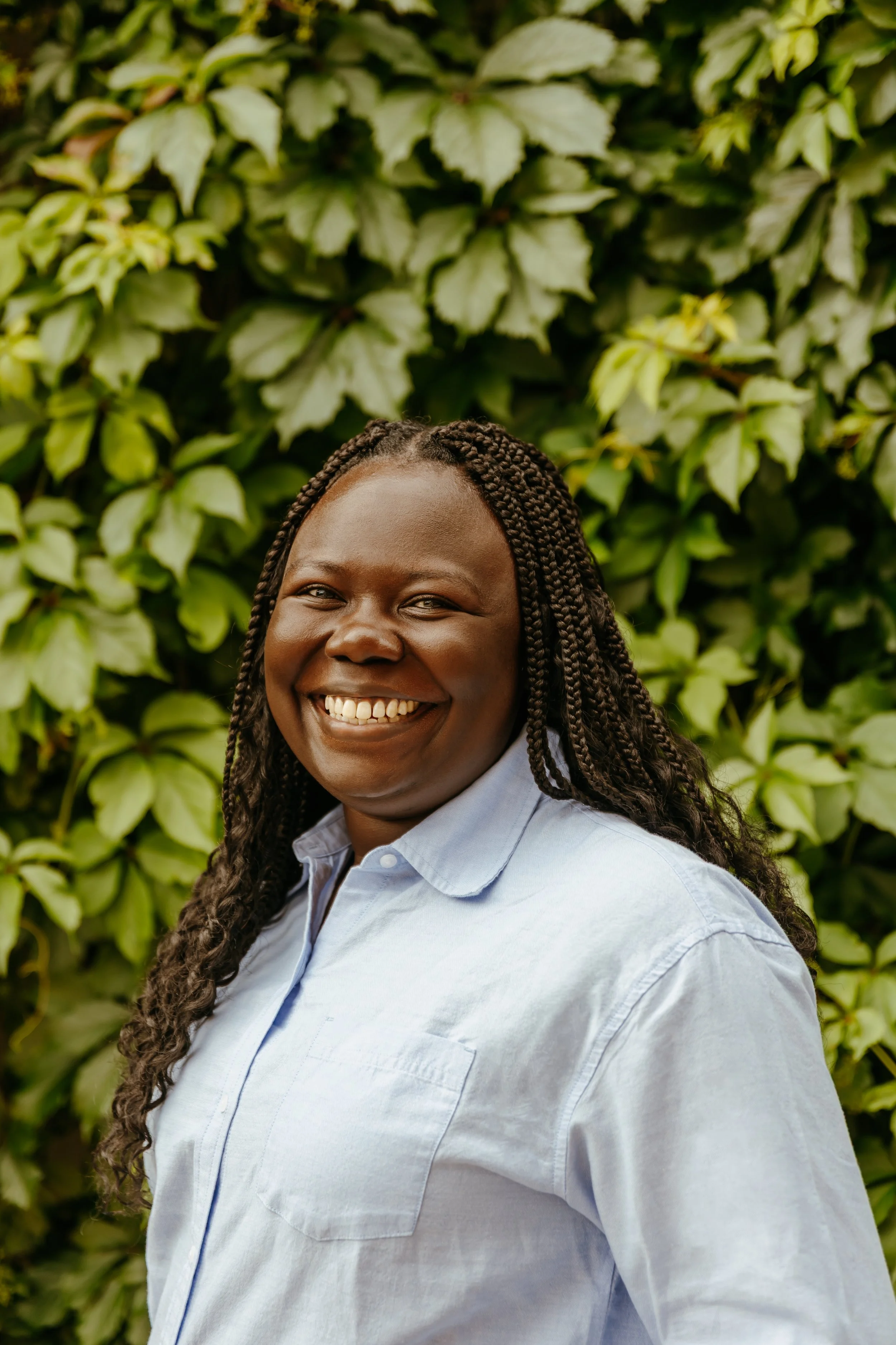 Smiling woman with braided hair wearing a light blue shirt standing in front of green leafy background.