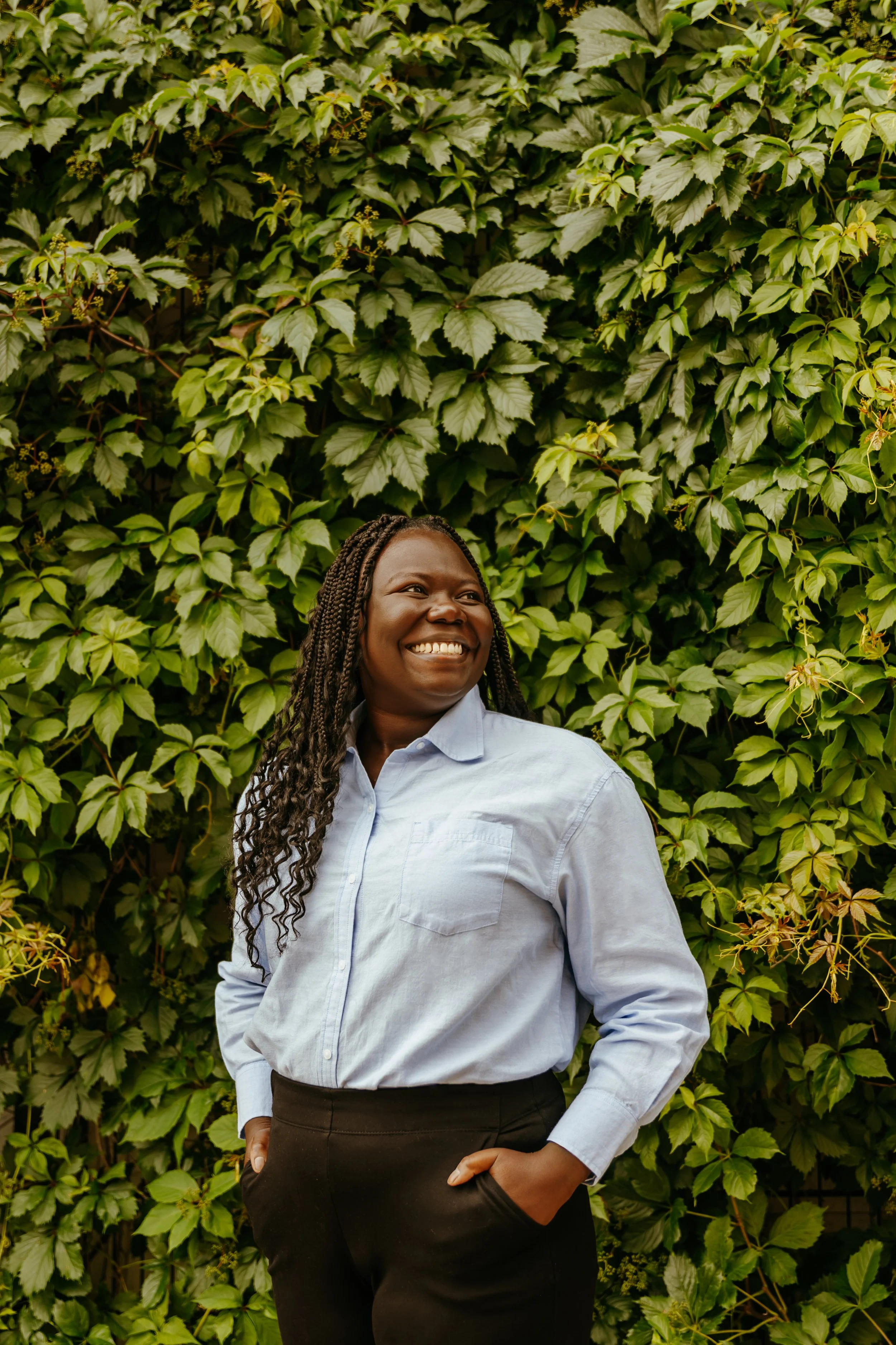 A smiling woman with long braided hair wearing a light blue button-up shirt and black pants stands in front of a lush green leafy wall, looking cheerful and relaxed.