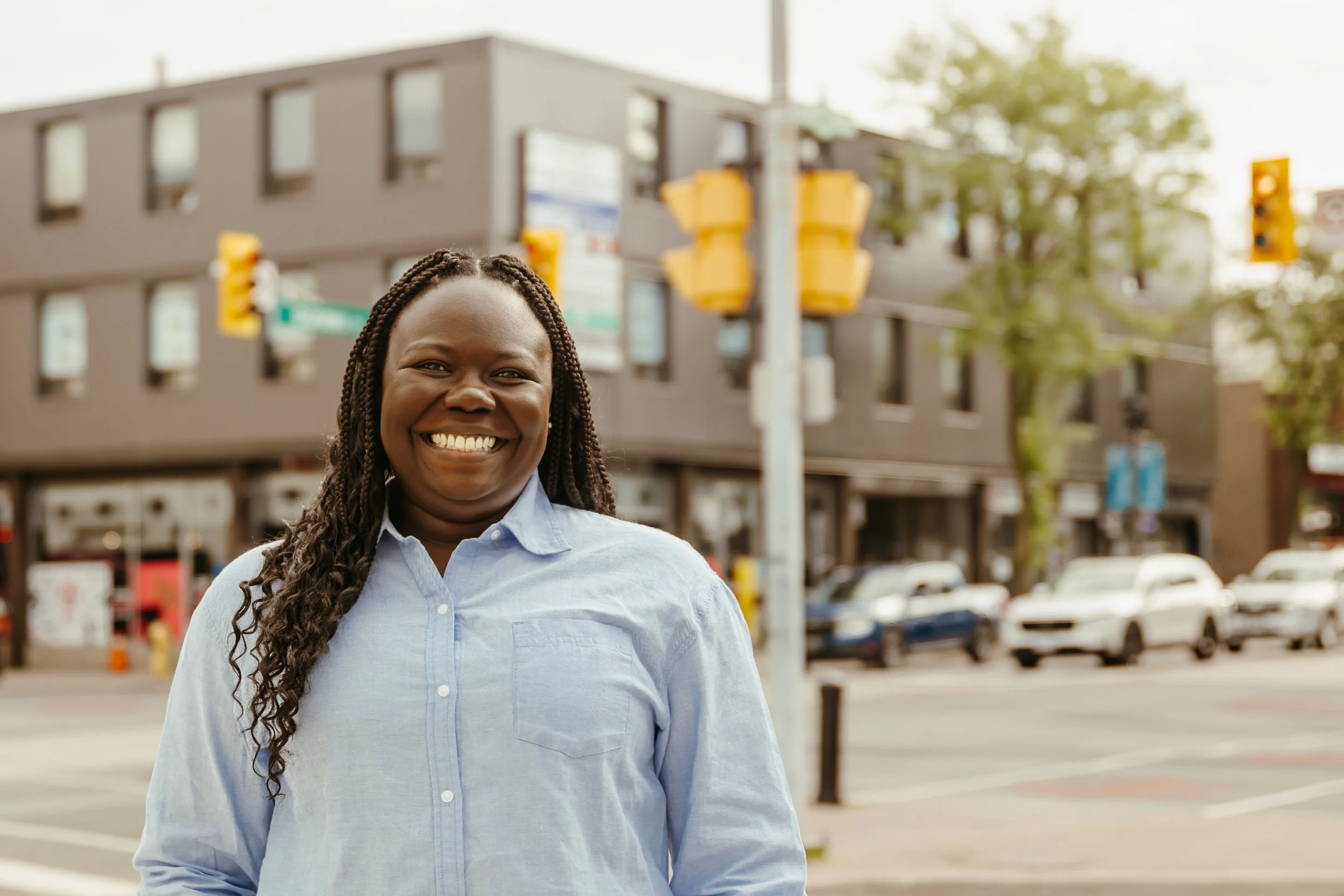 A woman with braided hair smiling outdoors in front of a city street scene with traffic lights, cars, and a modern building.