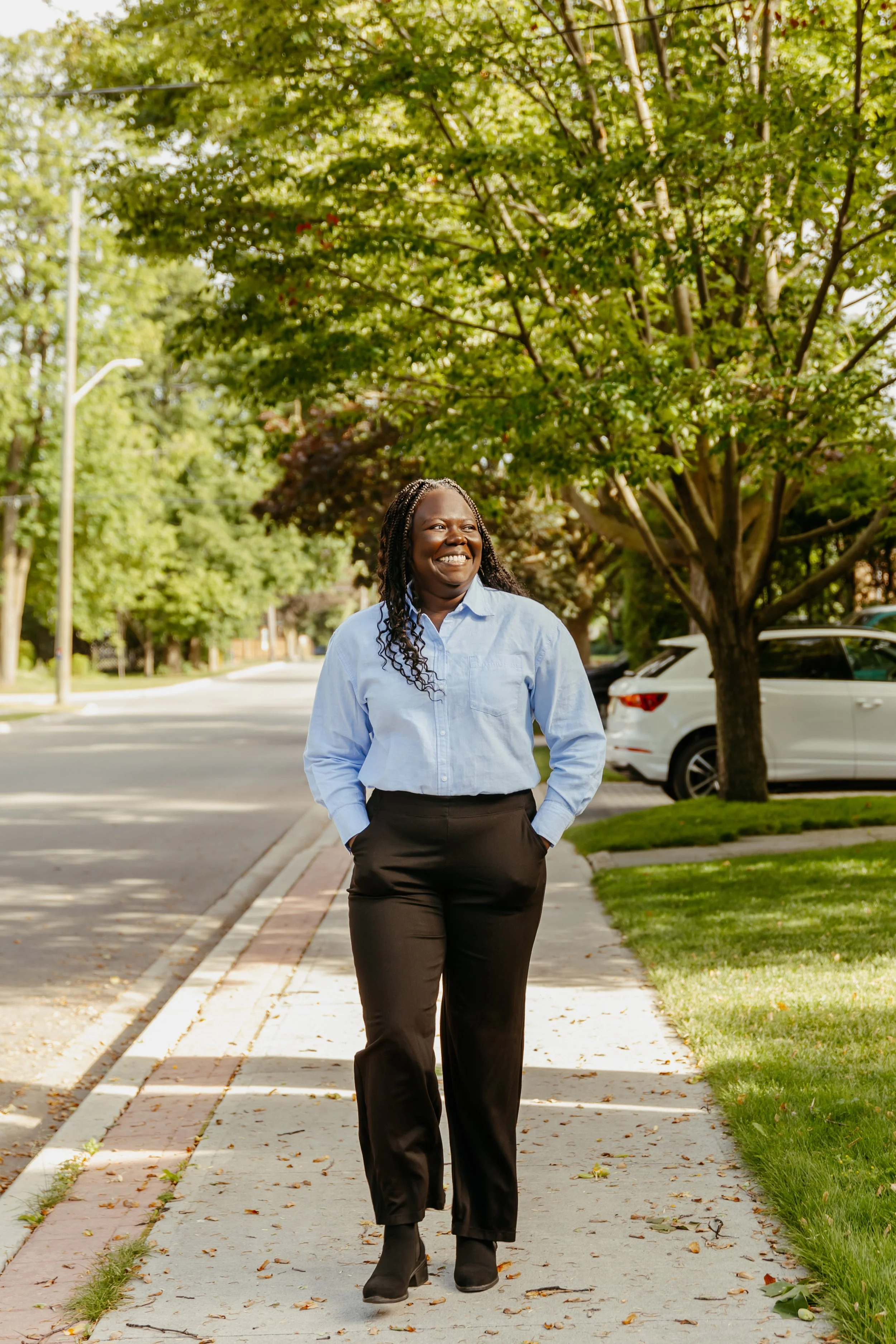 A woman in a light blue shirt and black pants walking on a sidewalk with trees and parked cars in the background, smiling.
