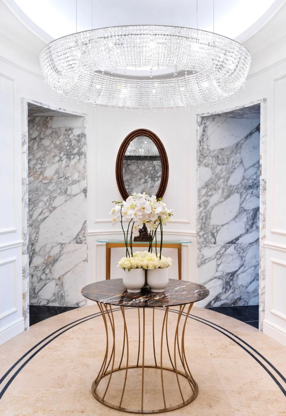 Elegant foyer with marble walls, a chandelier, a round table with a marble top, and vases of orchids and roses.