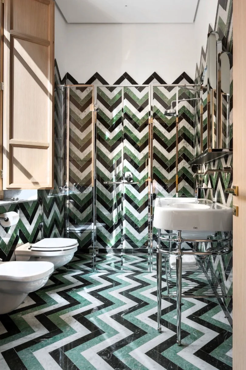Bathroom with black, white, green, and gray chevron patterned tiles on walls and floor, featuring a wall-mounted toilet, a glass shower enclosure, a white sink with chrome legs, and a wooden cabinet.