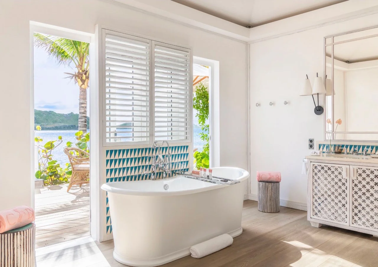 Bright bathroom with a view of a tropical outdoor scene including a palm tree and water. Features a white bathtub, white shutters, and decorative elements with blue geometric tiles.