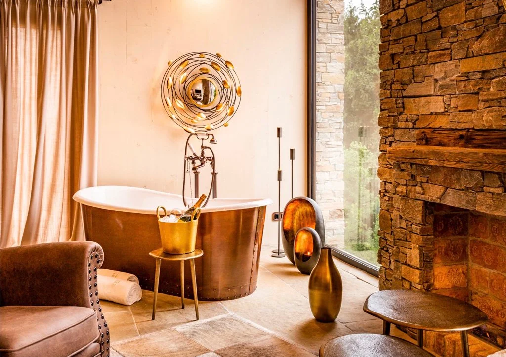Modern bathroom with a copper bathtub, tall candles, decorative vases, and a stone wall next to a large window.