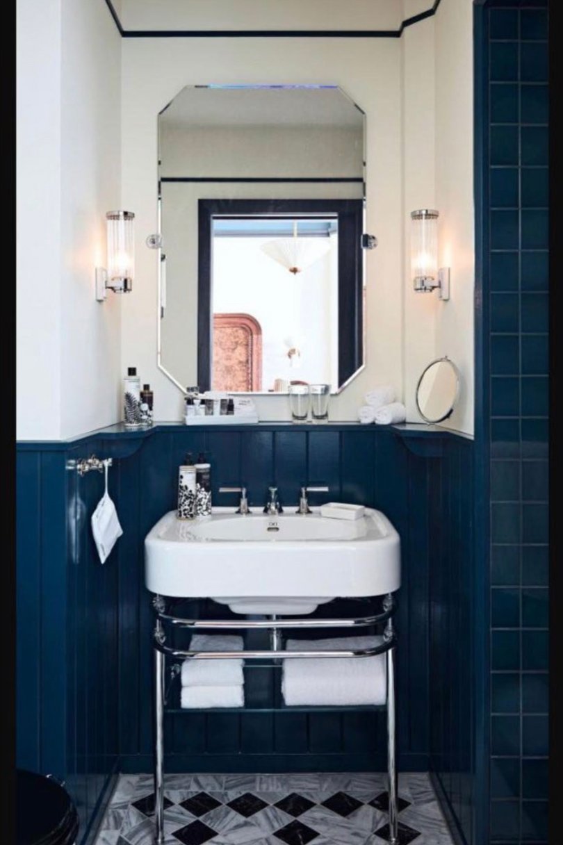 Bathroom with a white sink on a chrome stand, a mirror above it, blue wainscoting, wall-mounted lights, and a window reflecting a chandelier.