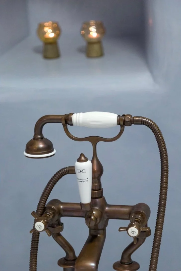 Vintage-style bathtub faucet with handheld shower head and two control knobs, mounted on a gray wall with two small gold-colored fixtures in the background.