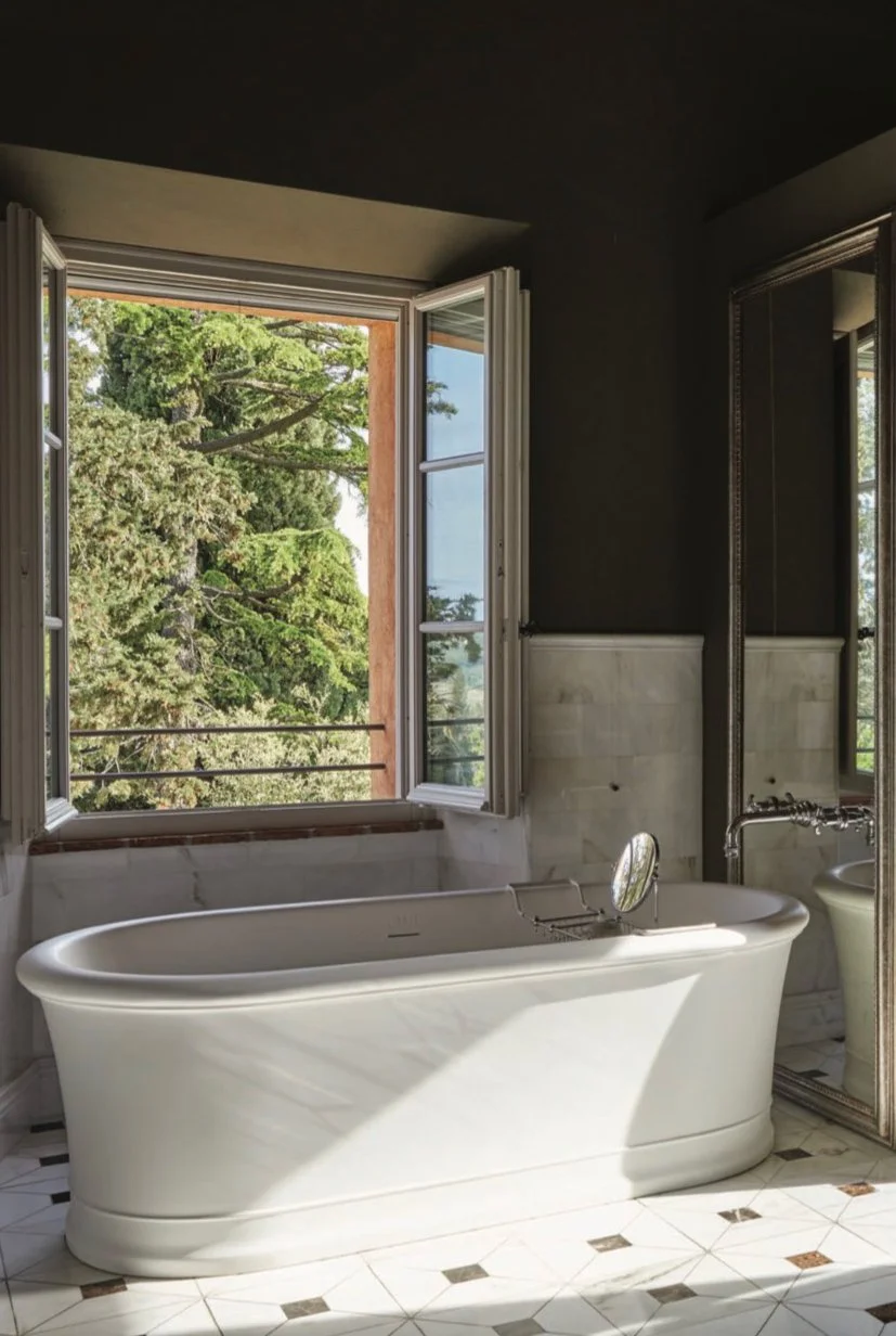 Bathroom with a white bathtub placed near a large open window showing green trees outside