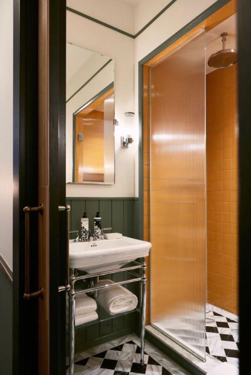 Small bathroom with a white sink, mirror, and a shower with orange tiles and a clear, ribbed shower door. There are towels under the sink and soap dispensers on the sink.