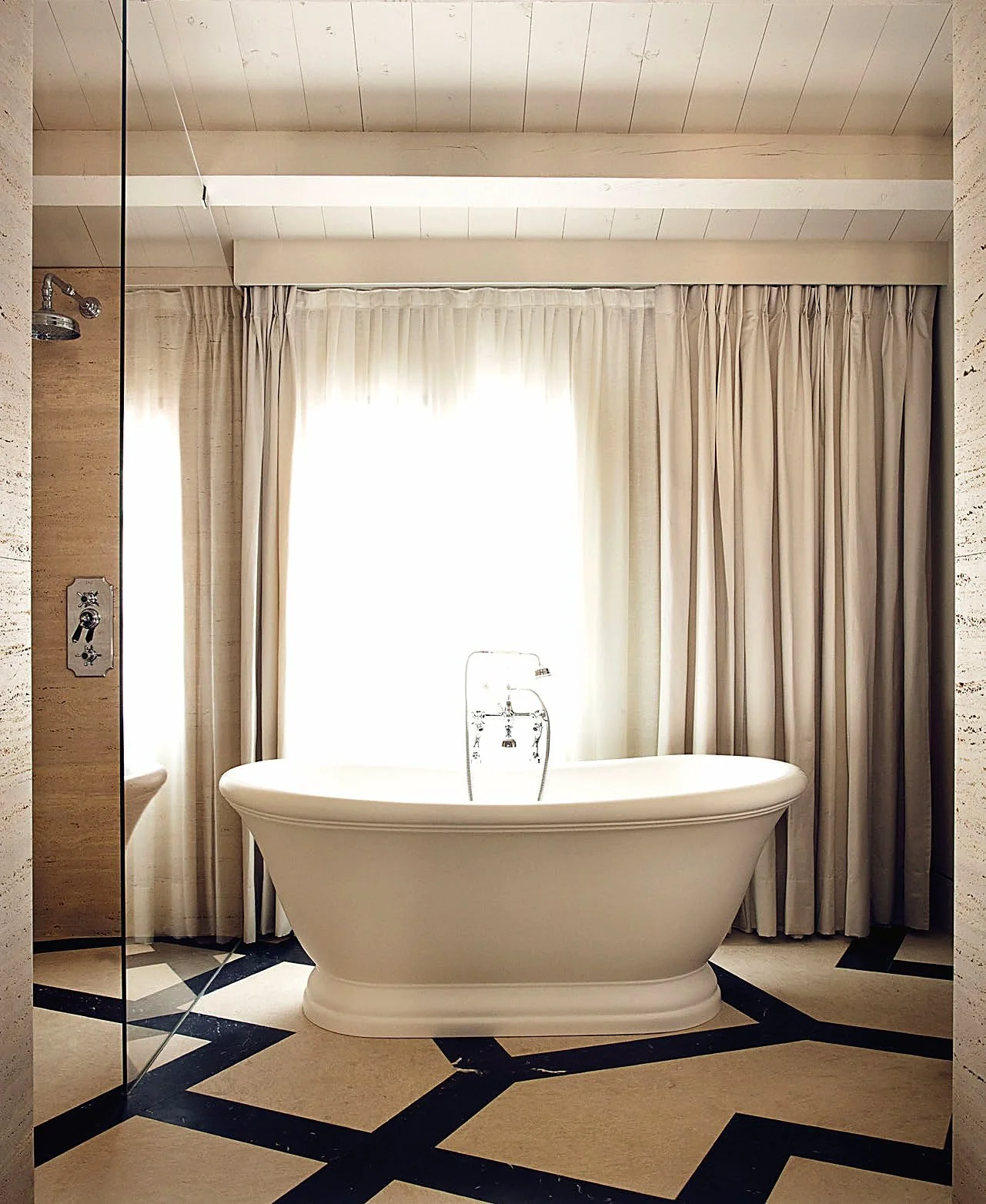 A white freestanding bathtub under a window with cream-colored curtains in a bathroom with beige walls and a patterned floor.