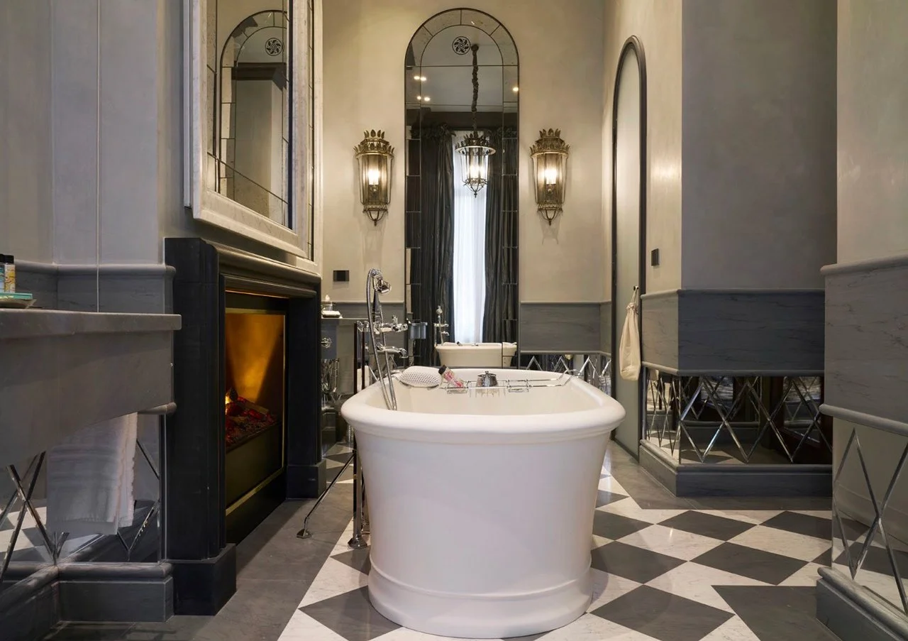 Elegant bathroom featuring a white clawfoot bathtub, black and white checkered floor, two wall-mounted lantern-style lights, a large mirror above the fireplace mantel, and tall windows with curtains, all in a sophisticated decor.