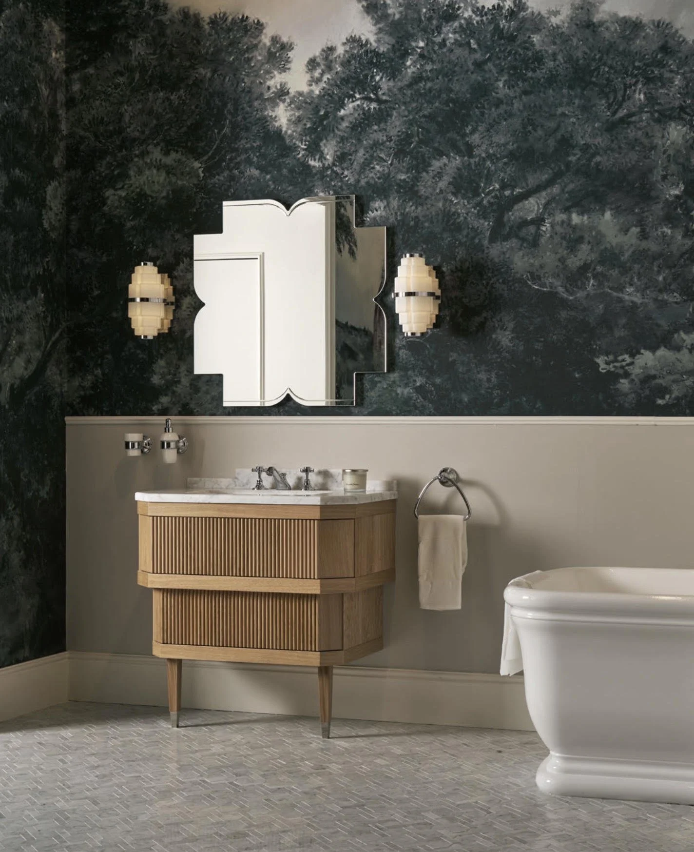 A bathroom decorated with a dark forest-themed wallpaper, a white framed mirror, two wall-mounted lamps, a modern wooden vanity with a marble countertop, a towel ring with a white towel, and a white vintage-style bathtub.