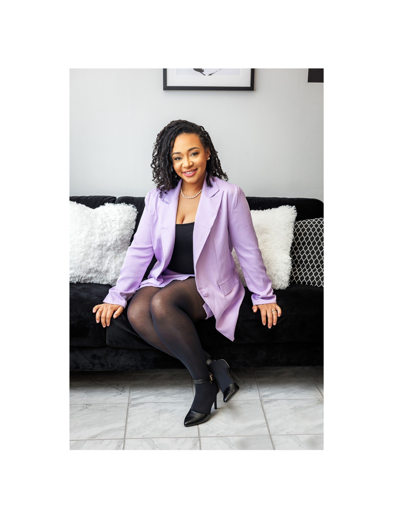 African American Realtor woman in NC with curly black hair and a warm smile sitting on a black couch with white and black pillows, wearing a lavender blazer, black tights, black high-heeled ankle boots, a pearl necklace, and earrings.