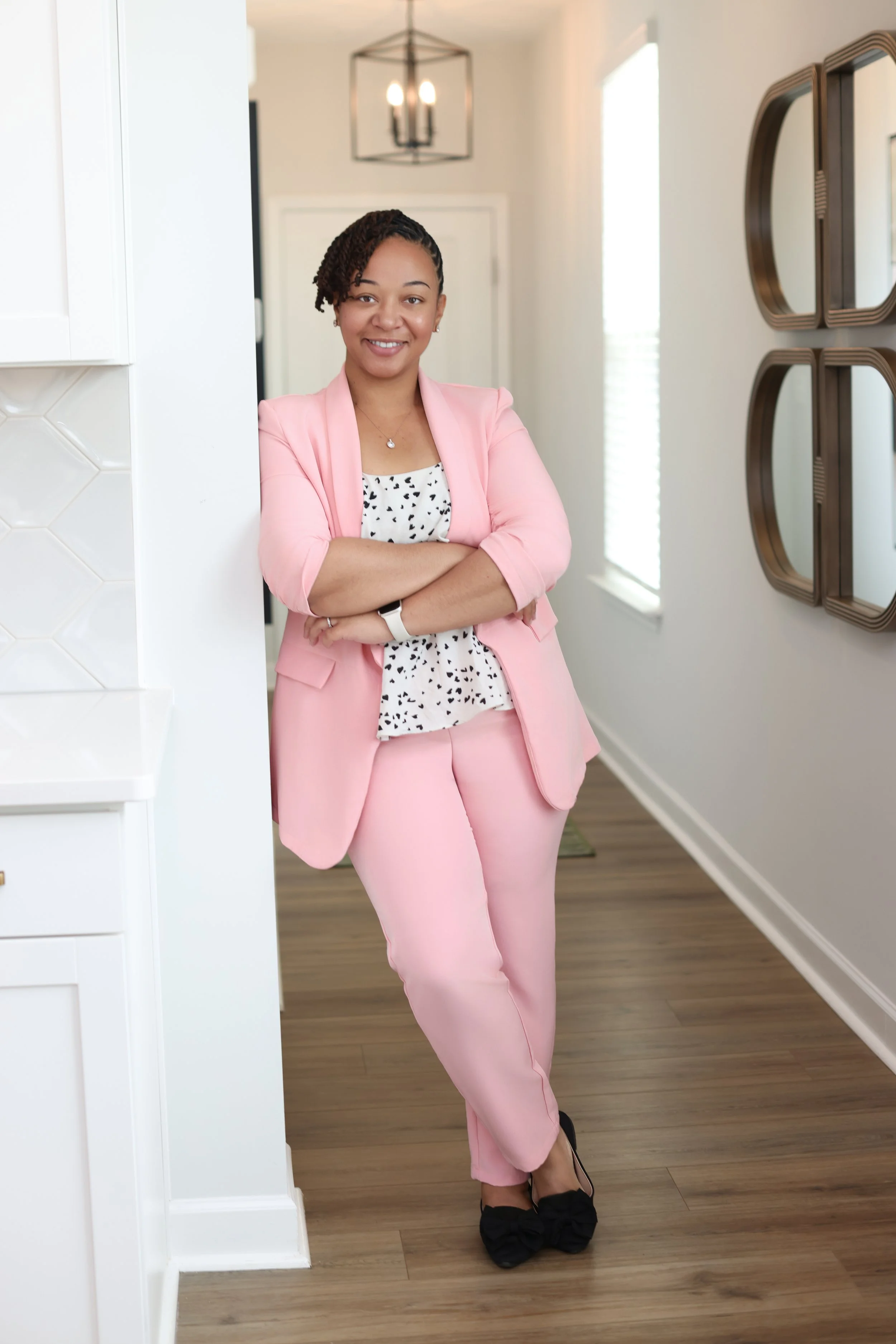North Carolina Realtor dressed in a pink blazer and matching pants, standing in a well-lit room with a modern decor, smiling with arms crossed.