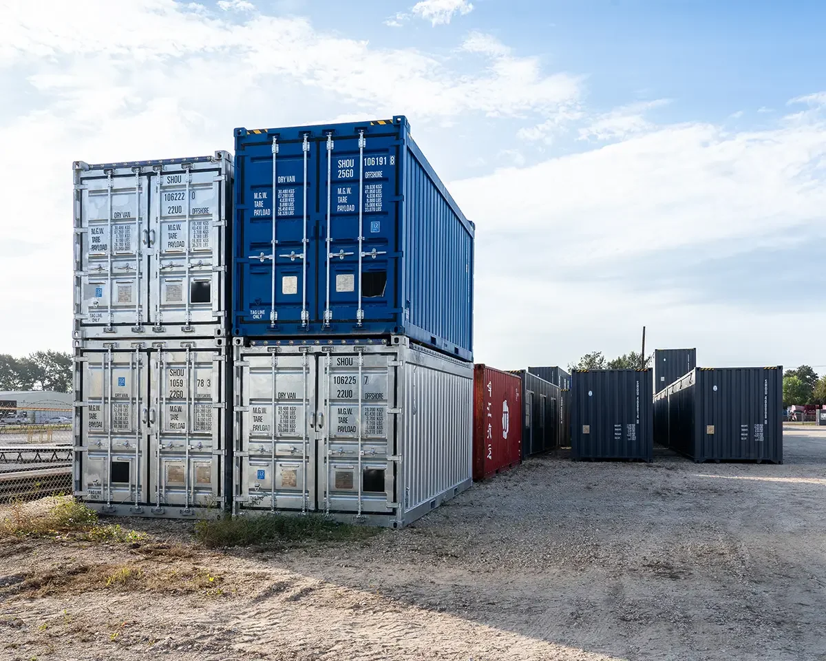 Stacked and lying shipping containers of various colors in an outdoor storage yard under a partly cloudy sky.