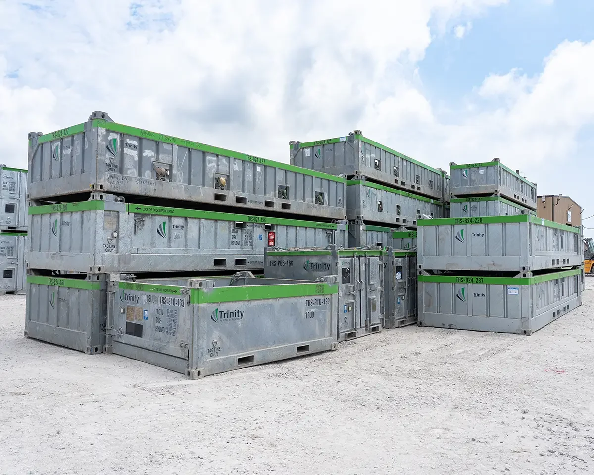 Multiple large gray storage containers with green borders stacked outdoors on a gravel surface, under a partly cloudy sky.