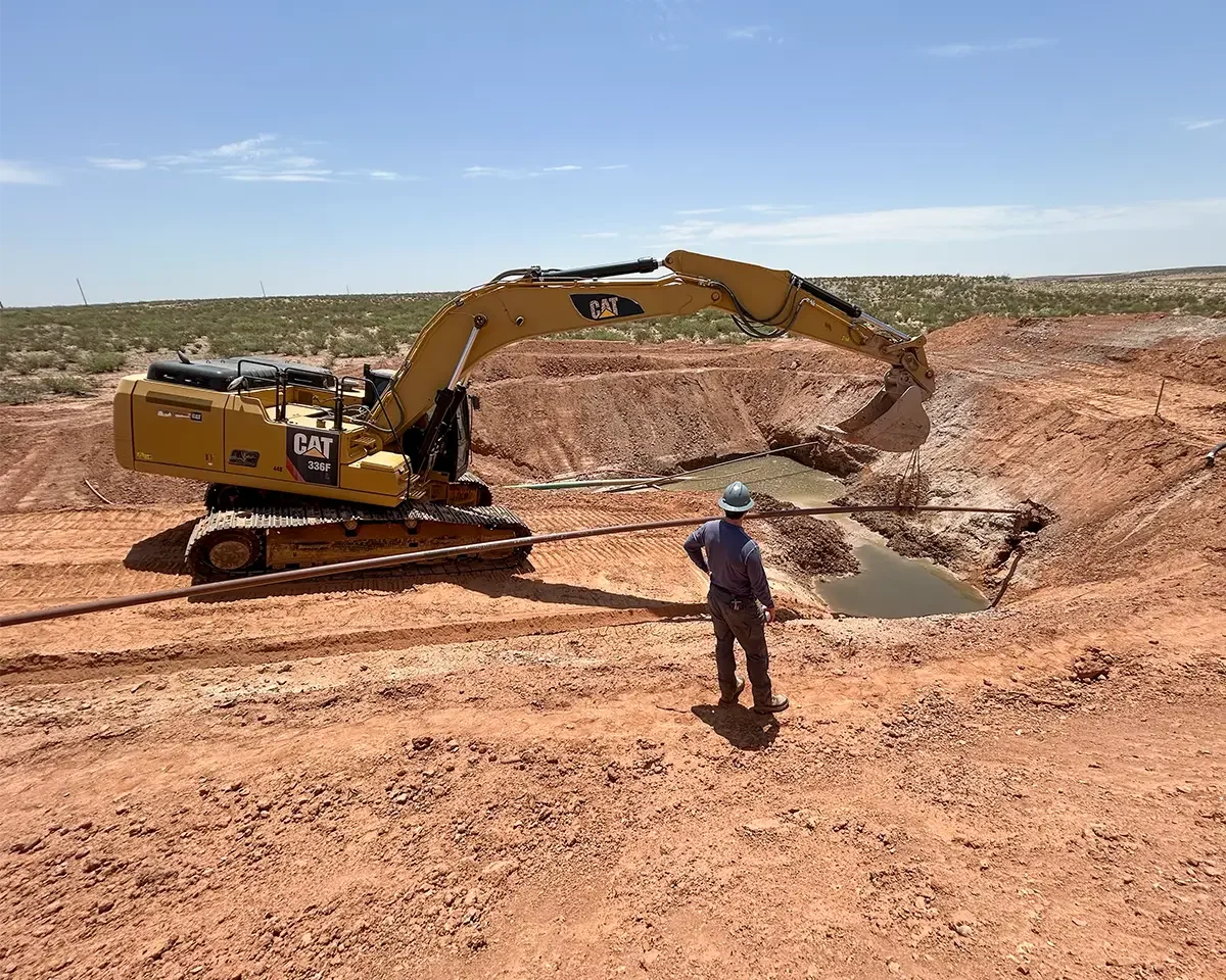 A construction worker in a hard hat observing a large yellow excavator digging a trench in a barren, reddish landscape on a sunny day.