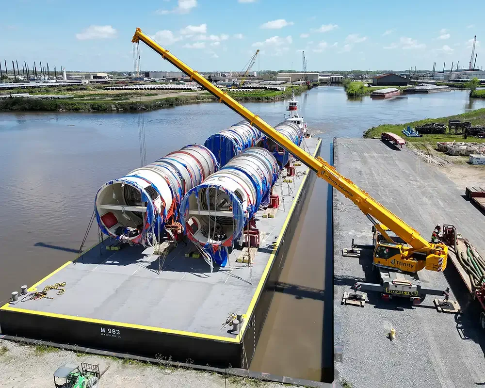 A barge is being loaded with large cylindrical sections, likely tunnel boring machine parts, by a yellow crane on a river. Industrial area with warehouses and cranes is visible in the background.