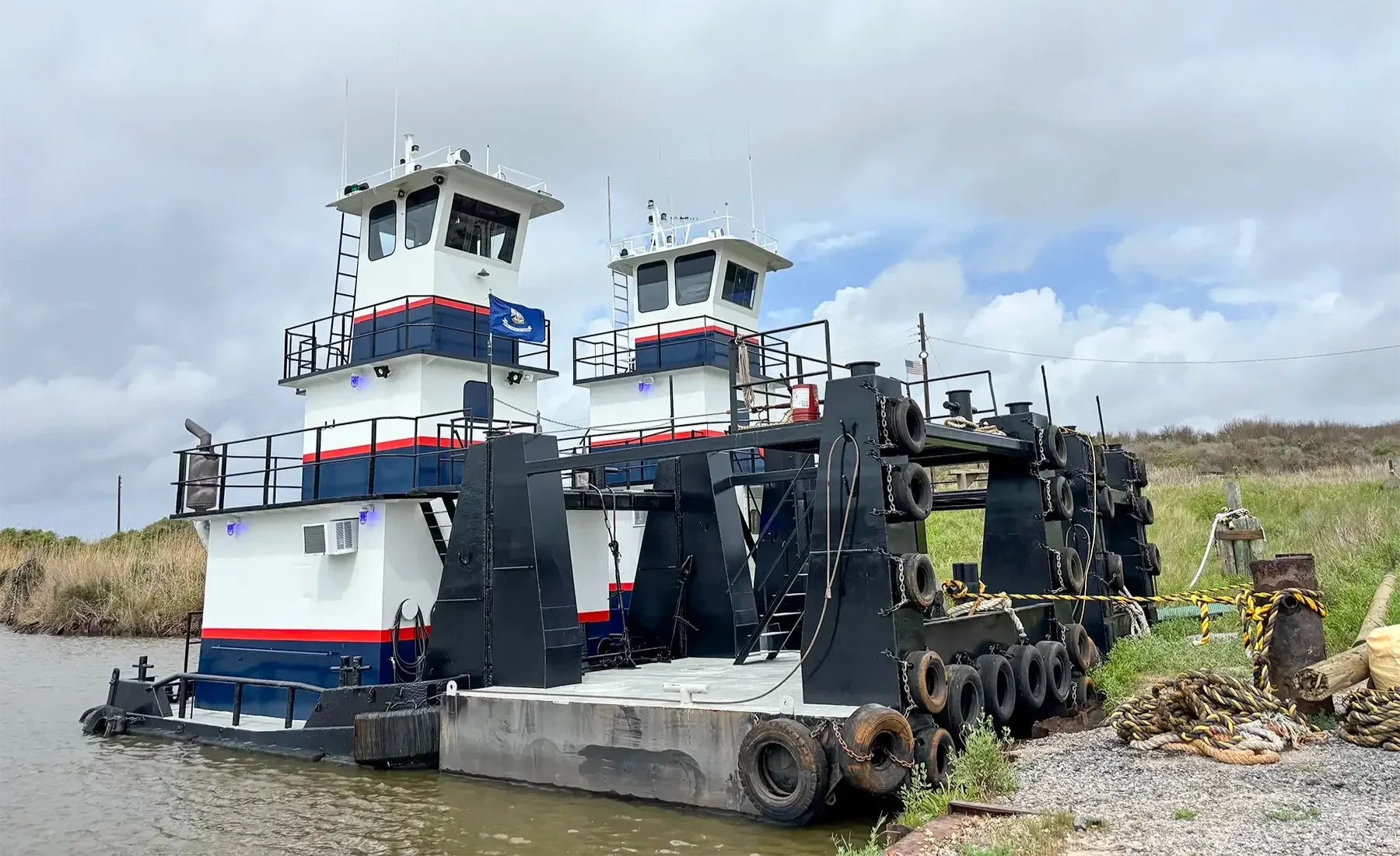 A large tugboat with white, blue, and red colors is docked near the shoreline, with a grassy area and cloudy sky in the background. The boat has two white cabins with black railings and an American flag. The hull is black, and thick black tires are attached along the side for bumper protection.