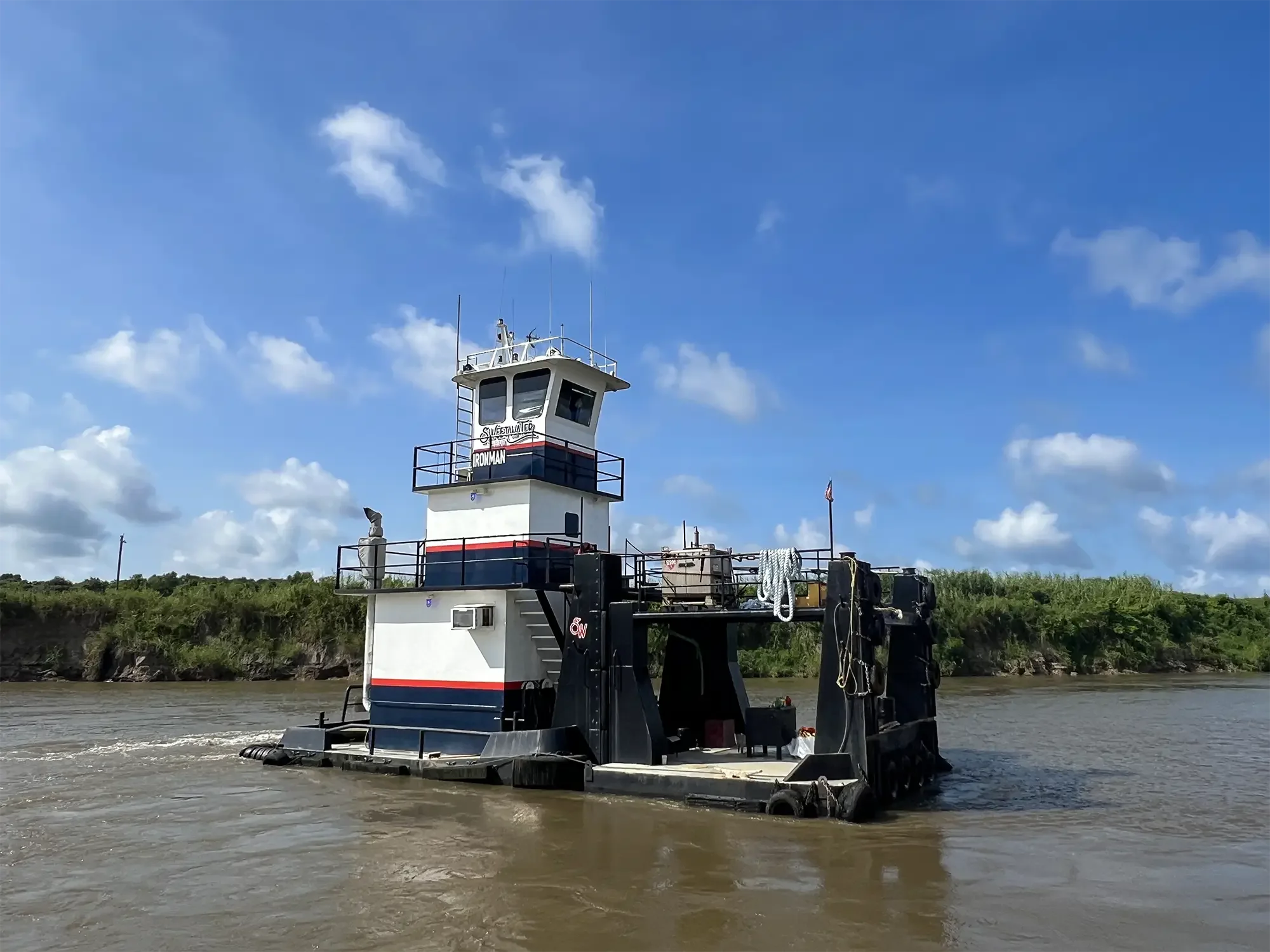 A black and white riverboat with a control tower on the river under a partly cloudy blue sky.