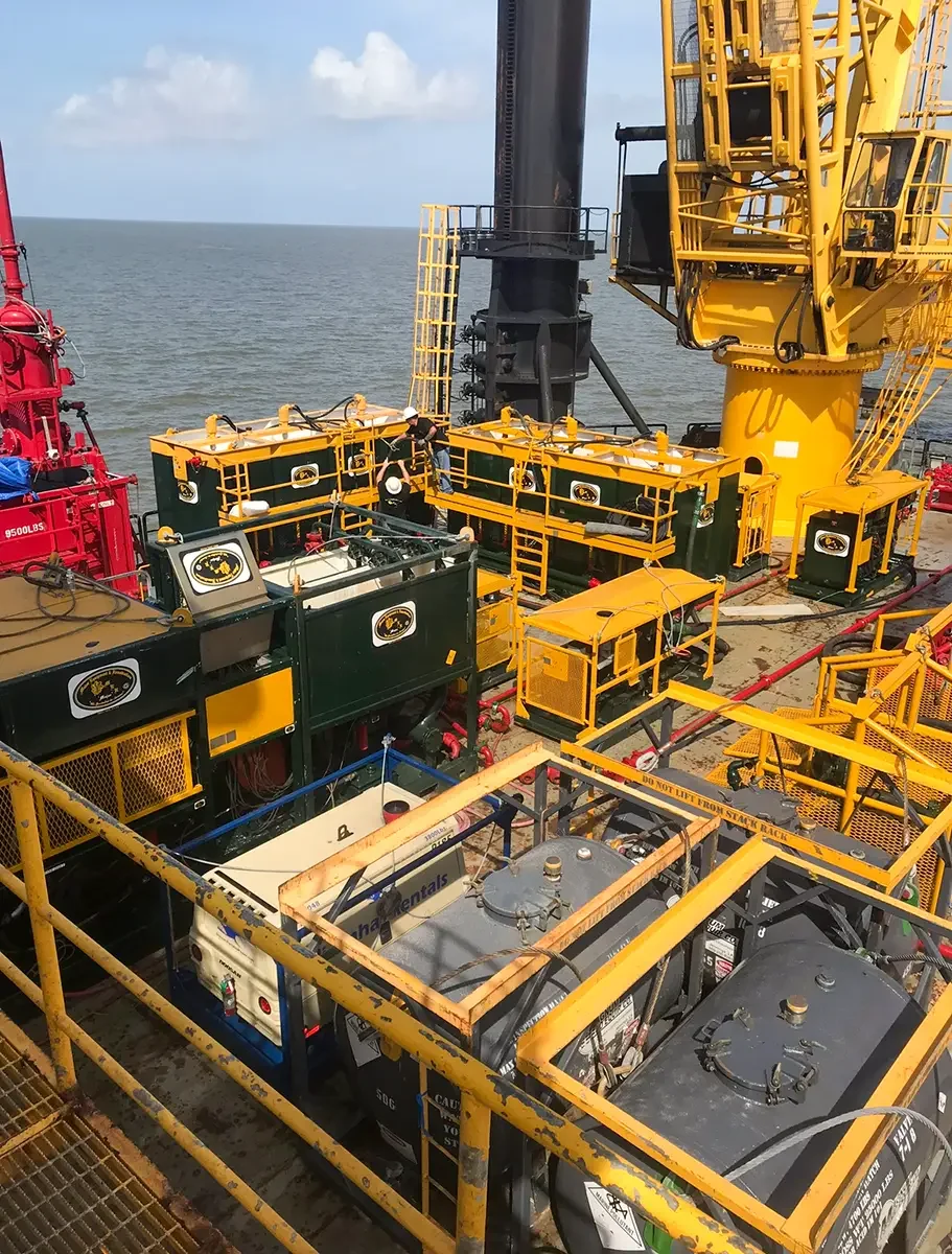 Close-up view of offshore oil drilling equipment on a platform, with yellow and green machinery and a large black drilling pipe extending into the ocean in the background.