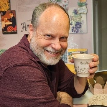 A man with gray hair and a beard smiling and holding a hot chocolate cup in a cozy chocolate shop.