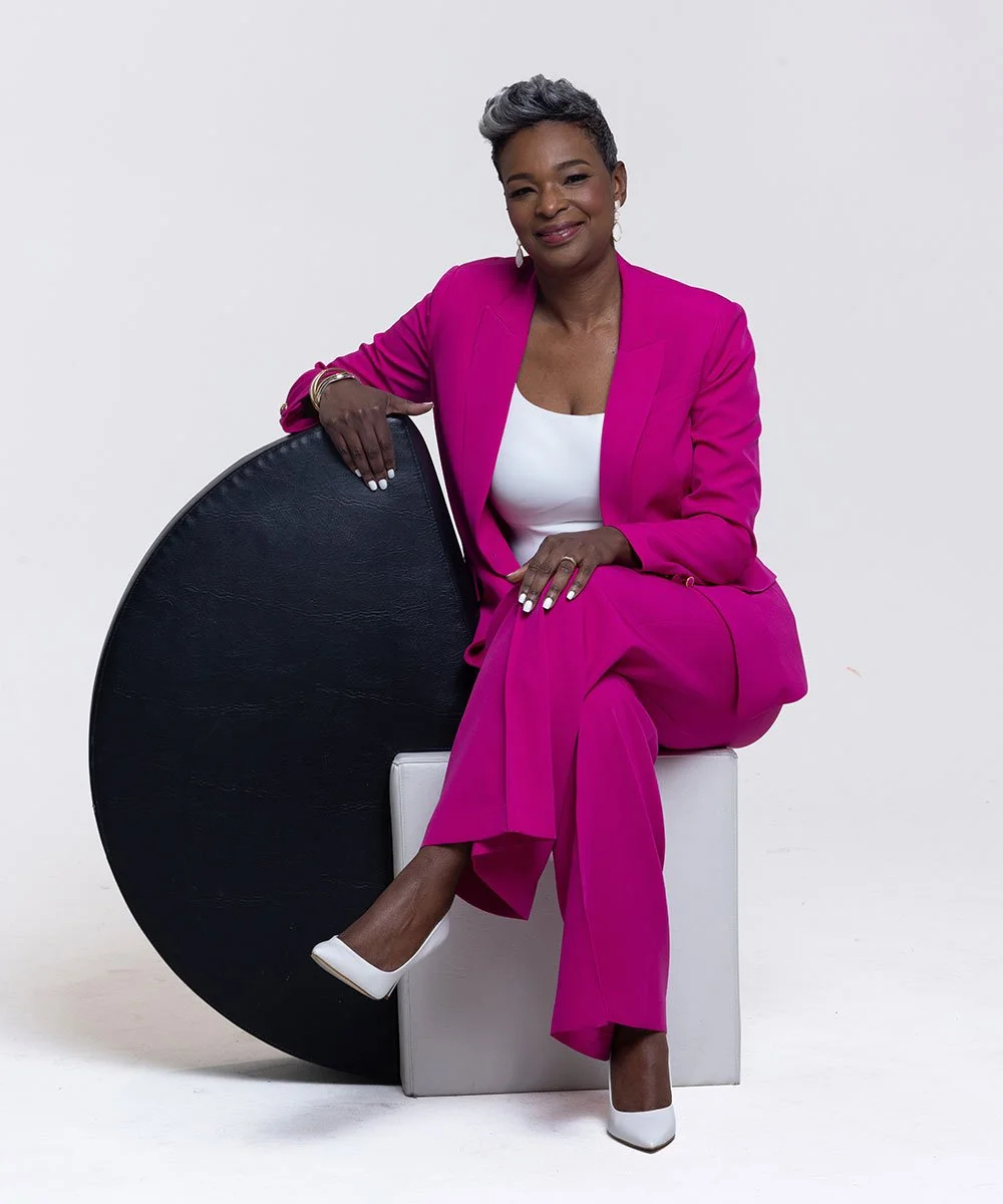A woman with short gray hair sitting on a white block, wearing a bright pink suit and white heels, smiling at the camera, with her left hand resting on her knee and her right hand on a black circular object.