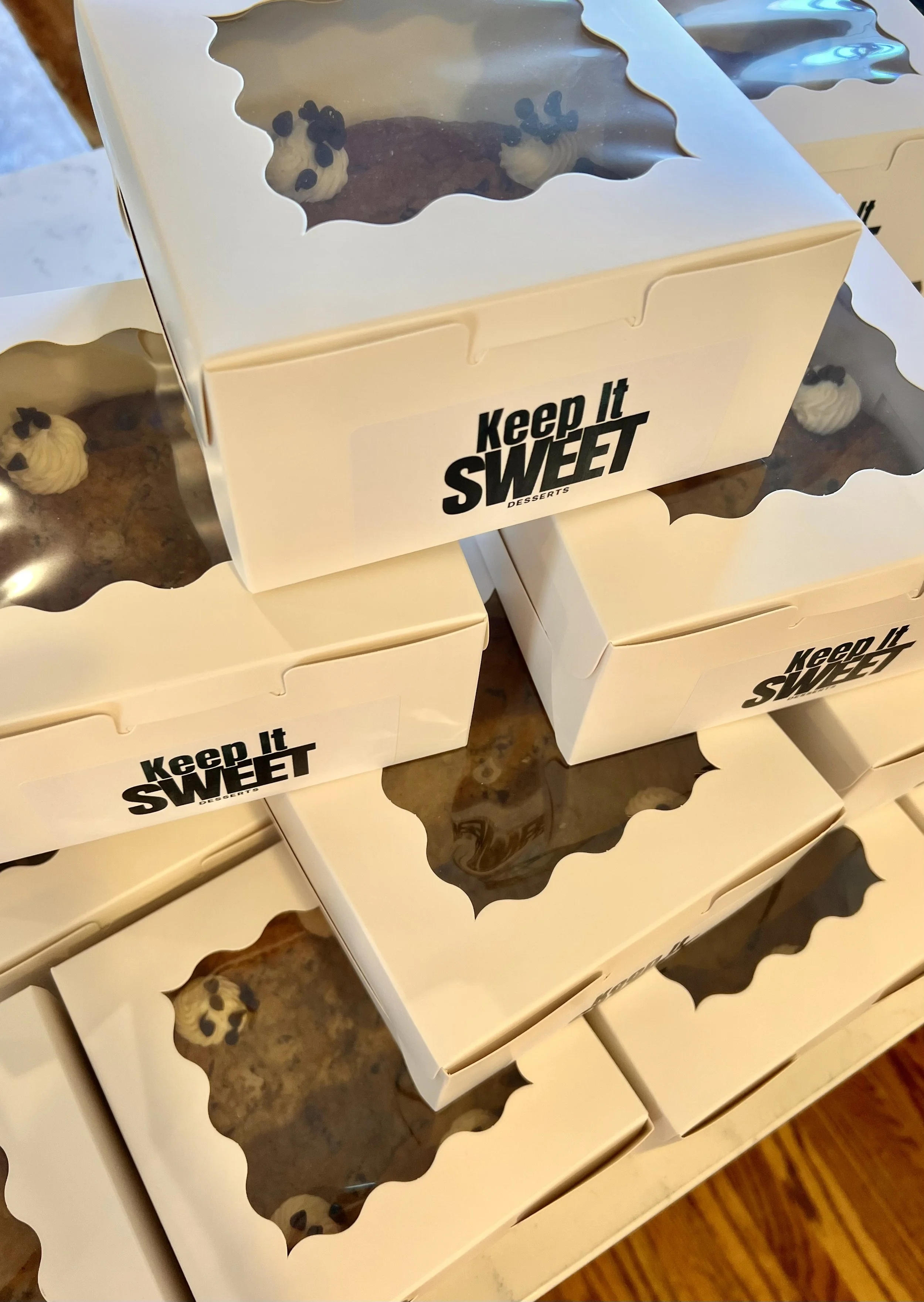Multiple white boxes of baked goods with transparent windows, labeled 'Keep It SWEET' stacked on a counter.