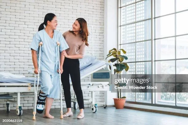 A woman in a hospital gown walking with crutches while supporting herself with another woman in a hospital room.