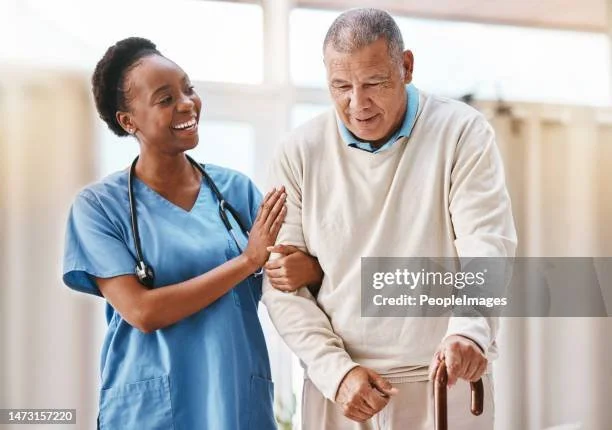 A nurse smiling and supporting an elderly man with a cane, in a healthcare setting.