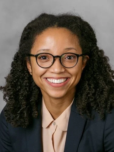 A woman with curly hair, glasses, and a nose ring, smiling in a professional setting.