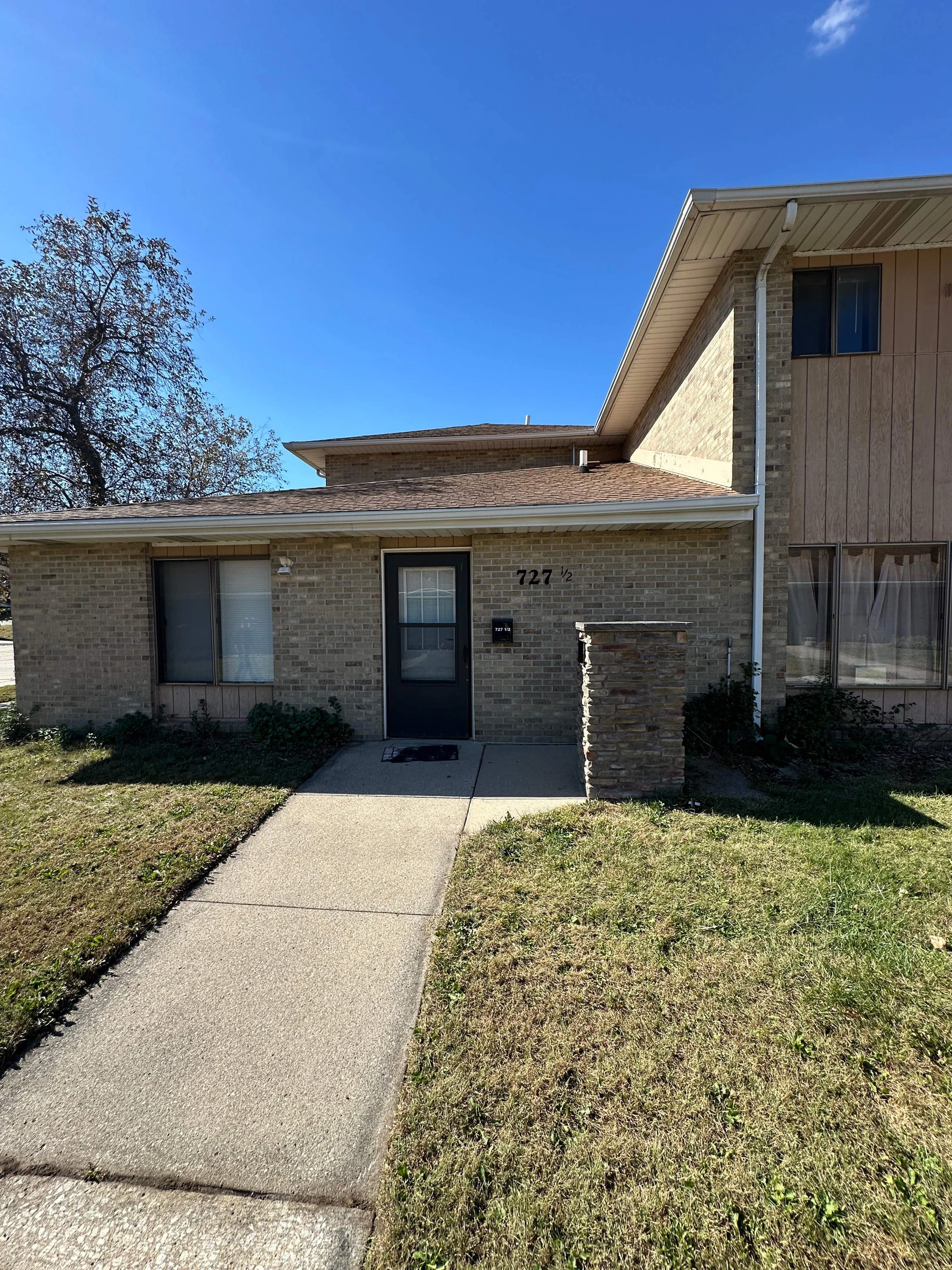 Front of a residential house with a concrete walkway leading to the front door, brick exterior, and lawn in the foreground.
