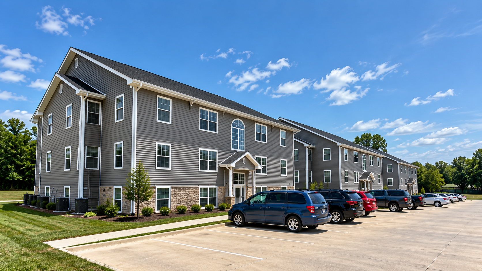 A multi-story apartment building with gray siding and white trim, surrounded by a well-kept lawn and small trees. There is a parking lot in front with multiple cars parked, under a bright blue sky with scattered clouds.