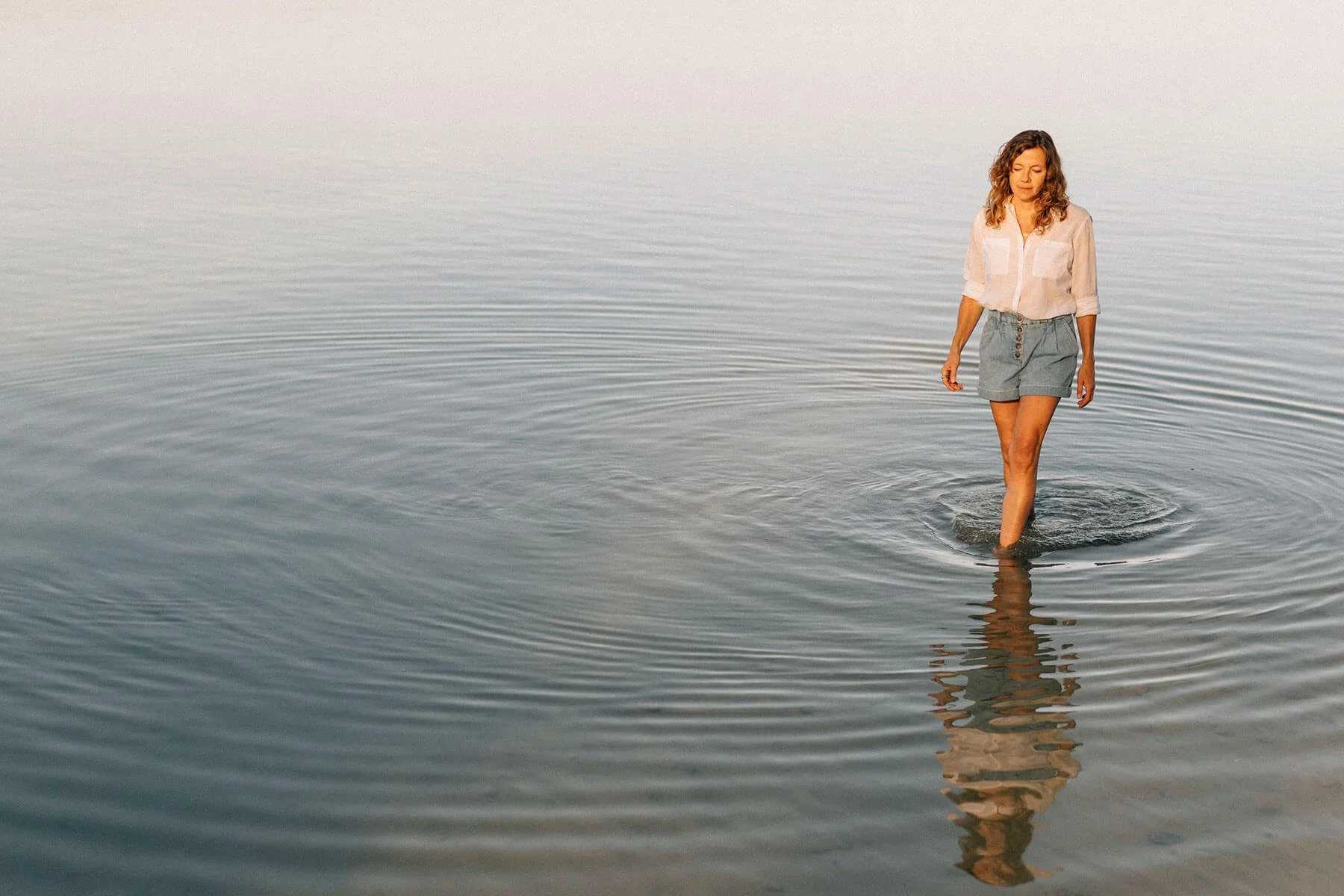 Anja Mai läuft barfuß durchs flache Wasser des Bodensees und blickt auf die Wasseroberfläche. Sie trägt eine weiße Bluse und eine kurze Jeanshose, ihre lockigen blonden Haare sind offen.