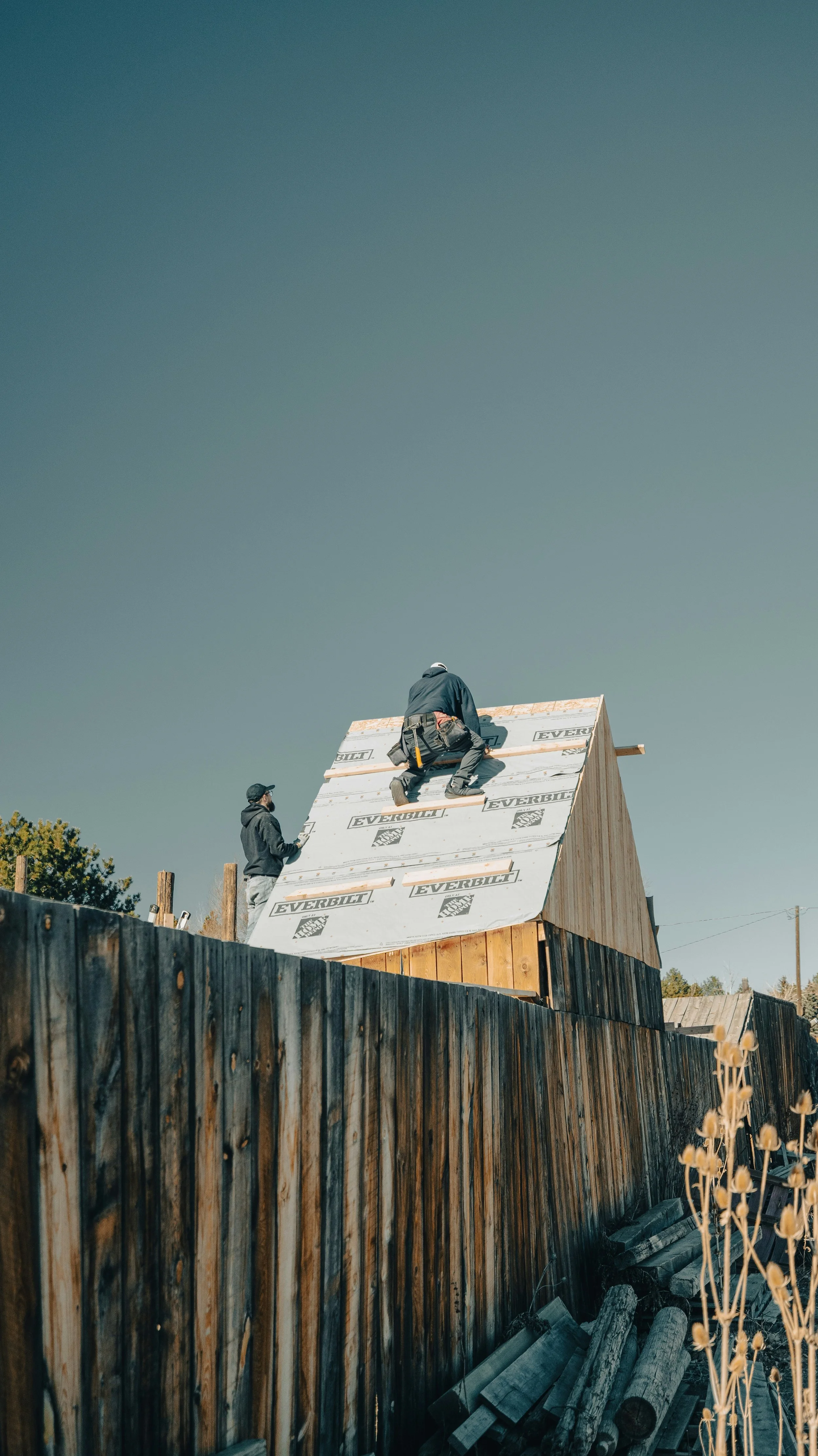 Two workers installing roofing on a small wooden house, one kneeling on the roof while the other stands nearby, with a tall wooden fence in the foreground and trees and clear sky in the background.
