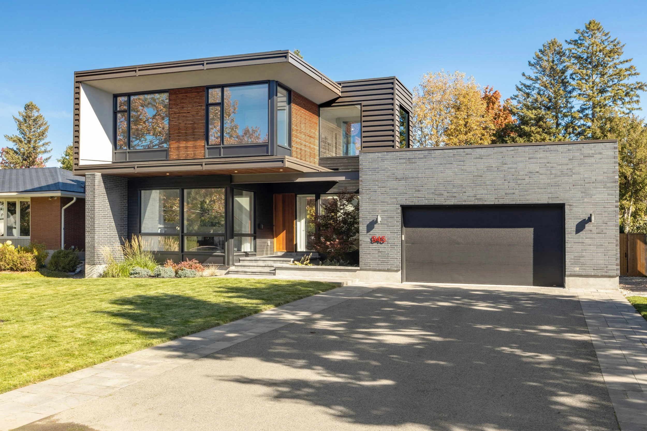 Modern two-story house with a gray brick exterior, large windows, and a garage, surrounded by a green lawn and trees with autumn foliage.