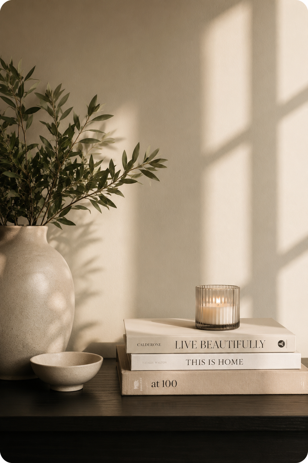 A beige vase with green leafy branches, a small matching bowl, a candle in a glass holder, and three books stacked on a dark surface, with sunlight casting shadows on a white wall.