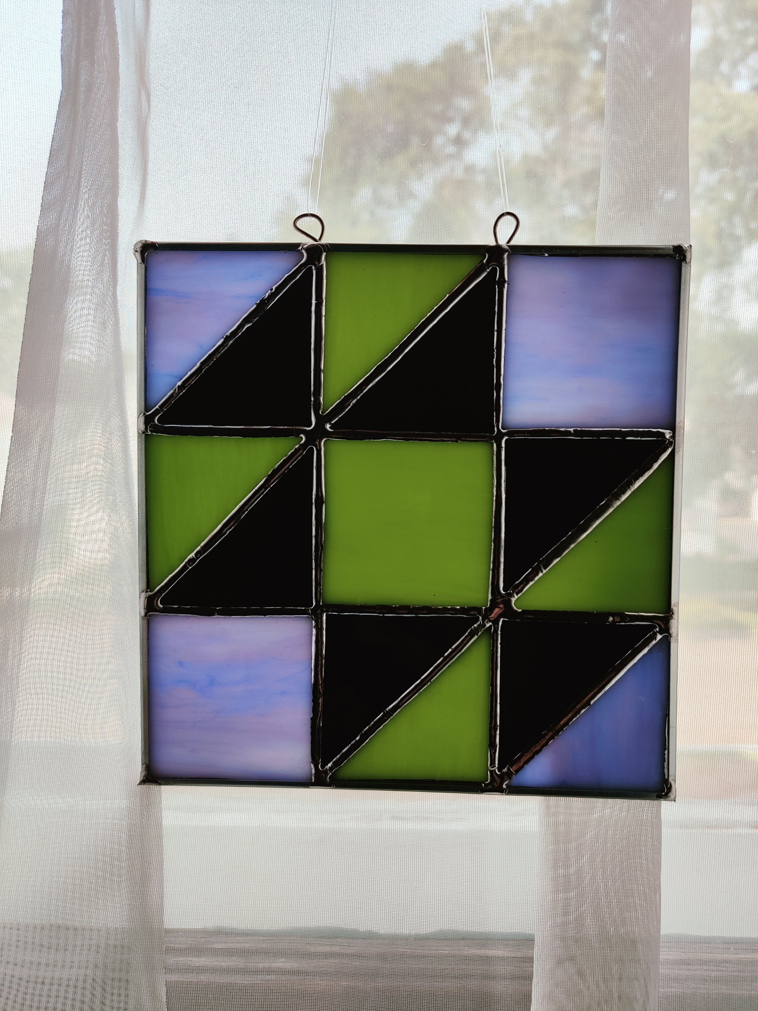 Stained glass window with geometric pattern in green, black, and shades of blue and purple, hanging in front of a sheer curtain with trees visible outside.