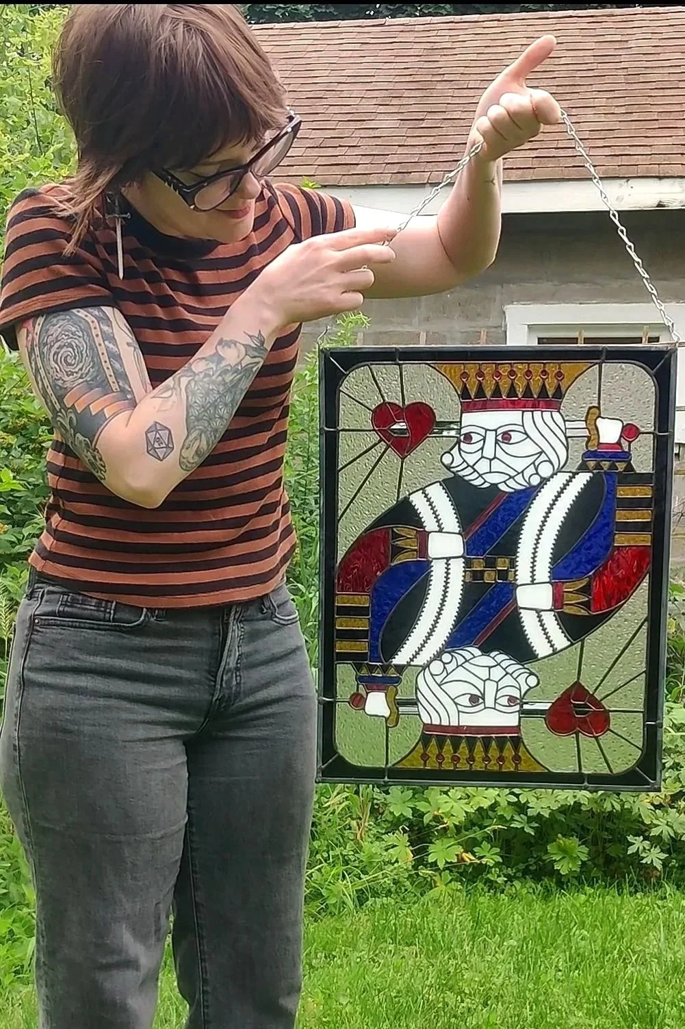 A woman with tattoos, wearing glasses and a striped shirt, holding a stained glass panel of a king playing card in a garden.