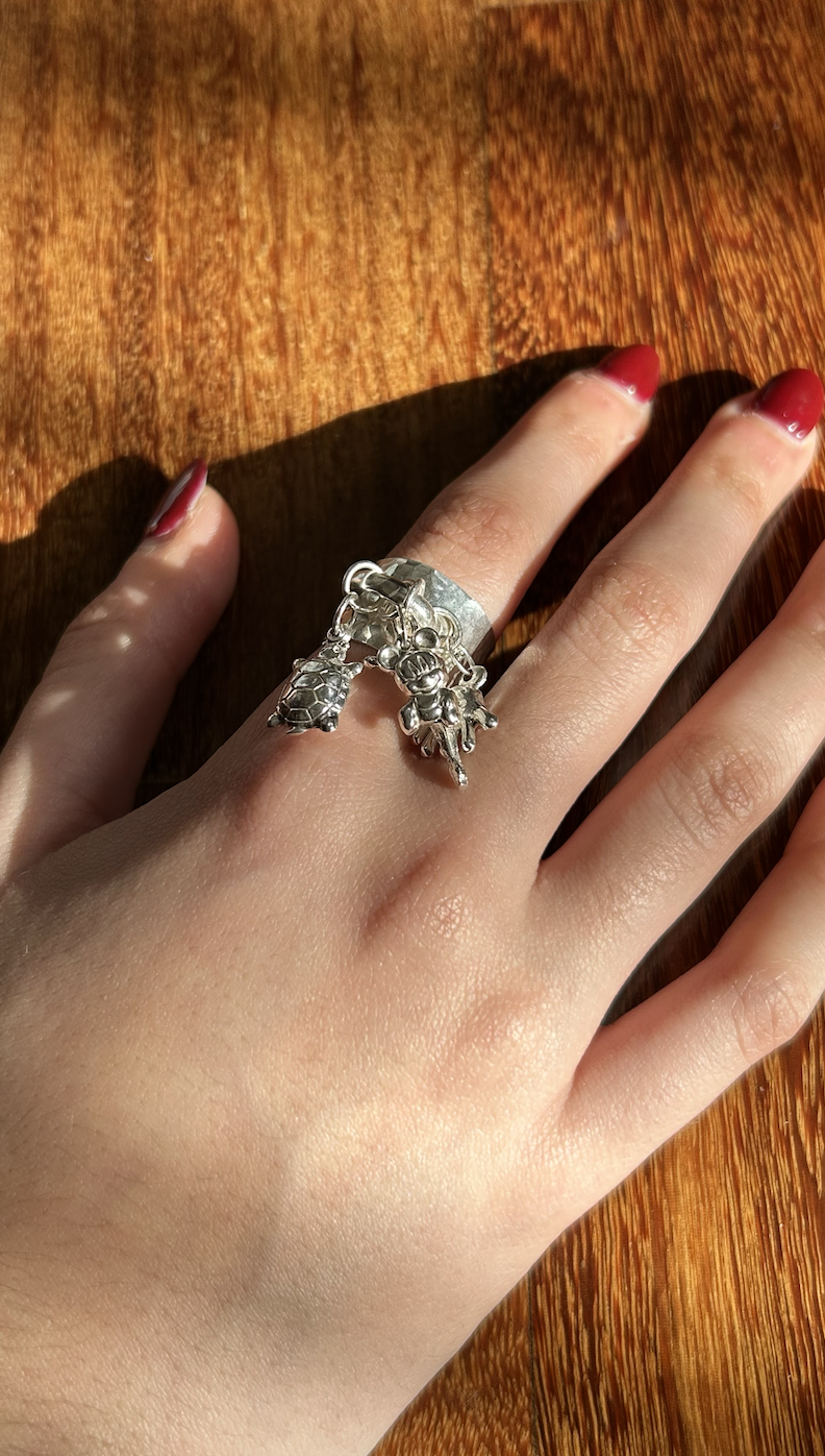 A person's hand resting on a wooden surface, wearing a large silver ring with multiple small charms hanging from it, including a bee, a star, and other shapes. The person has red nail polish.