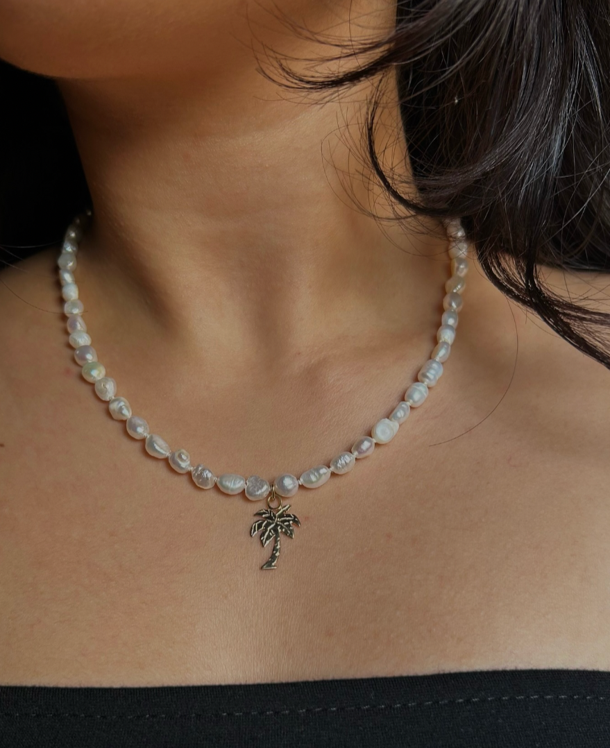 A woman is wearing a pearl necklace with a palm tree pendant. The image is a close-up of her neck and part of her shoulder, showing her dark hair and black clothing.