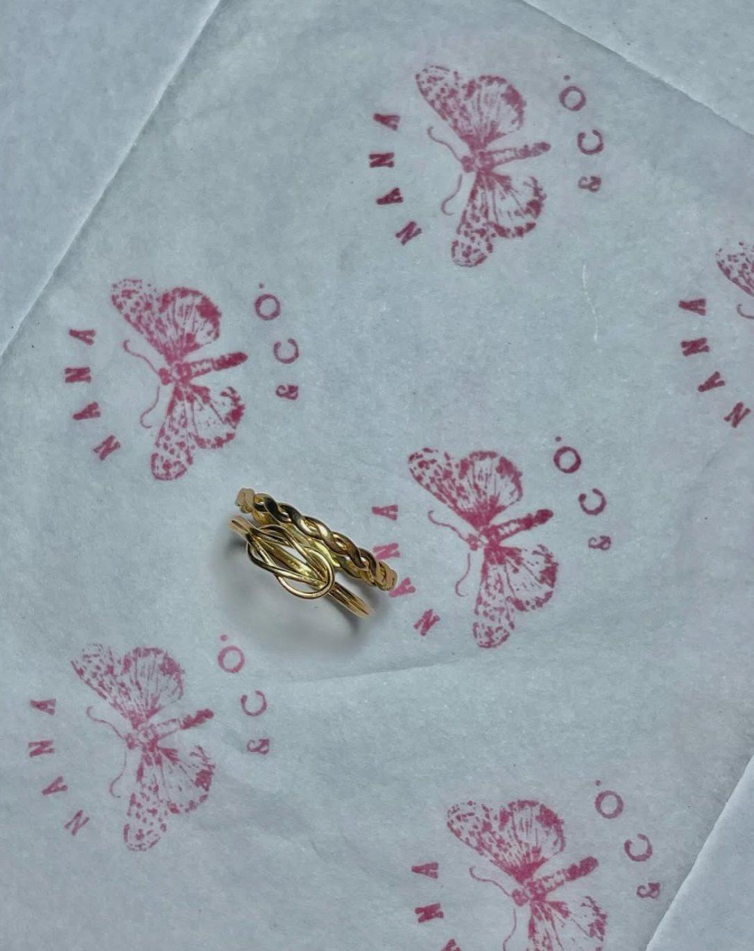 Gold rings placed on branded tissue paper with butterfly logo and the words 'HANA SCARO' printed in pink.