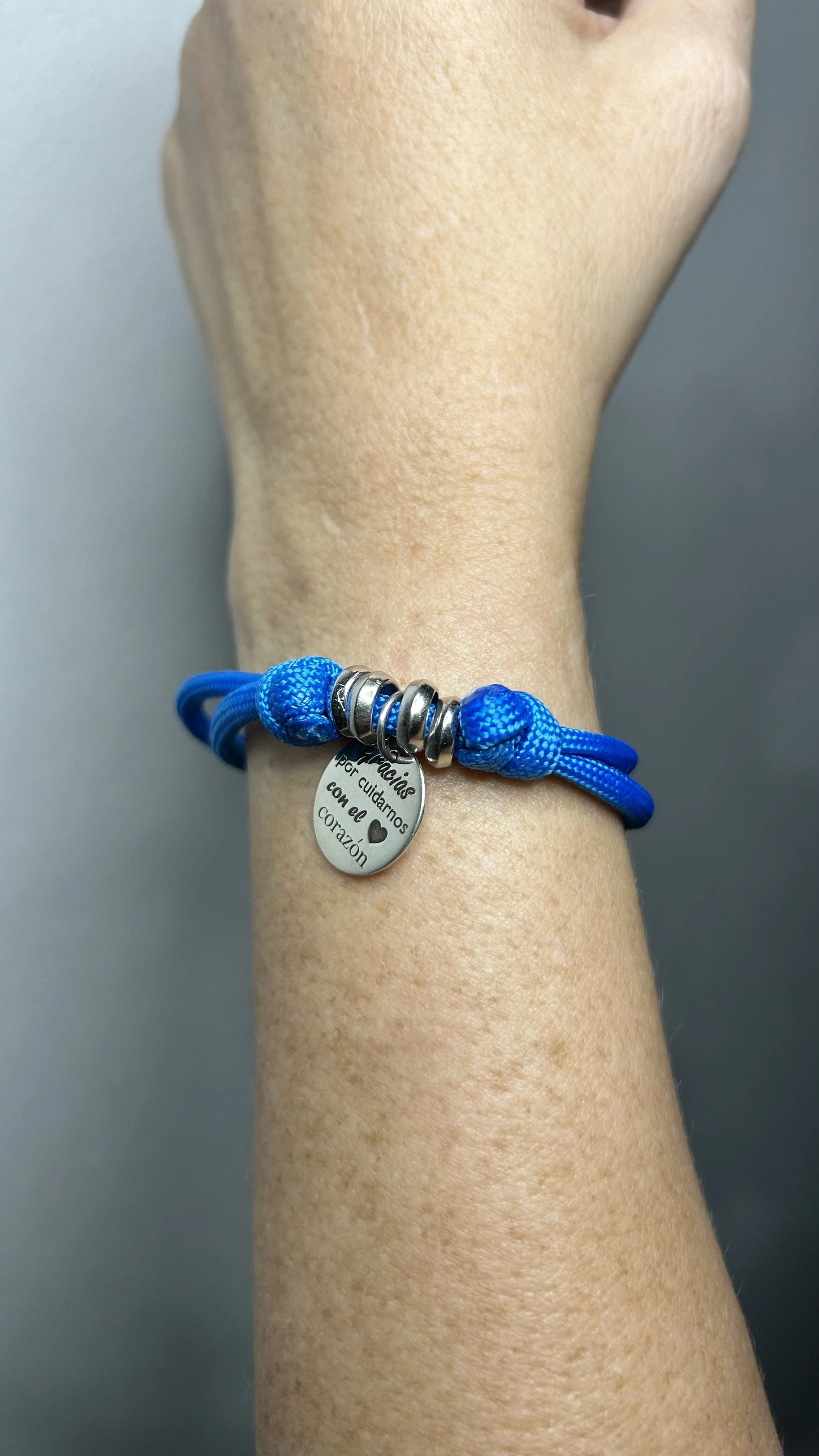 A wrist wearing a blue braided bracelet with silver beads and a small oval metal tag with Spanish text and a heart symbol.
