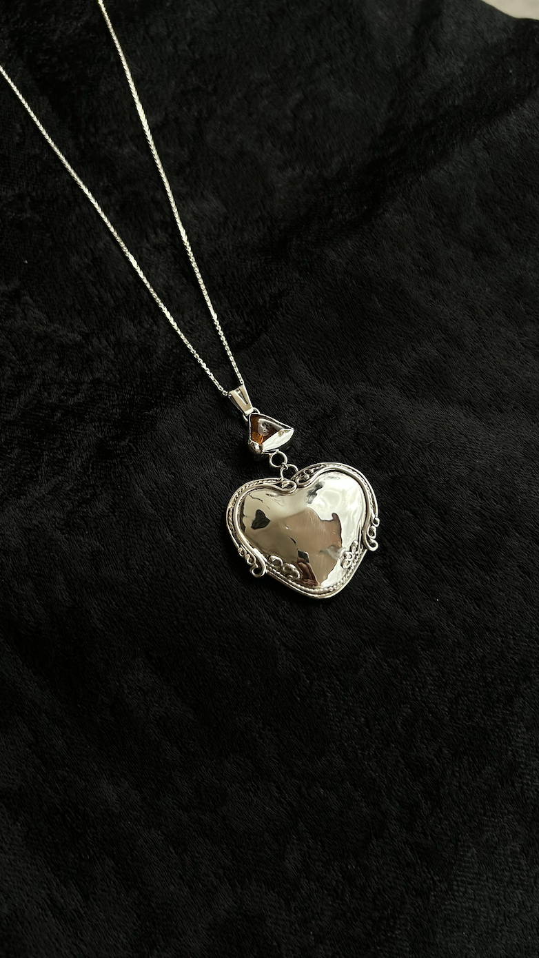 Silver heart-shaped pendant necklace with a decorative border on a black fabric background.
