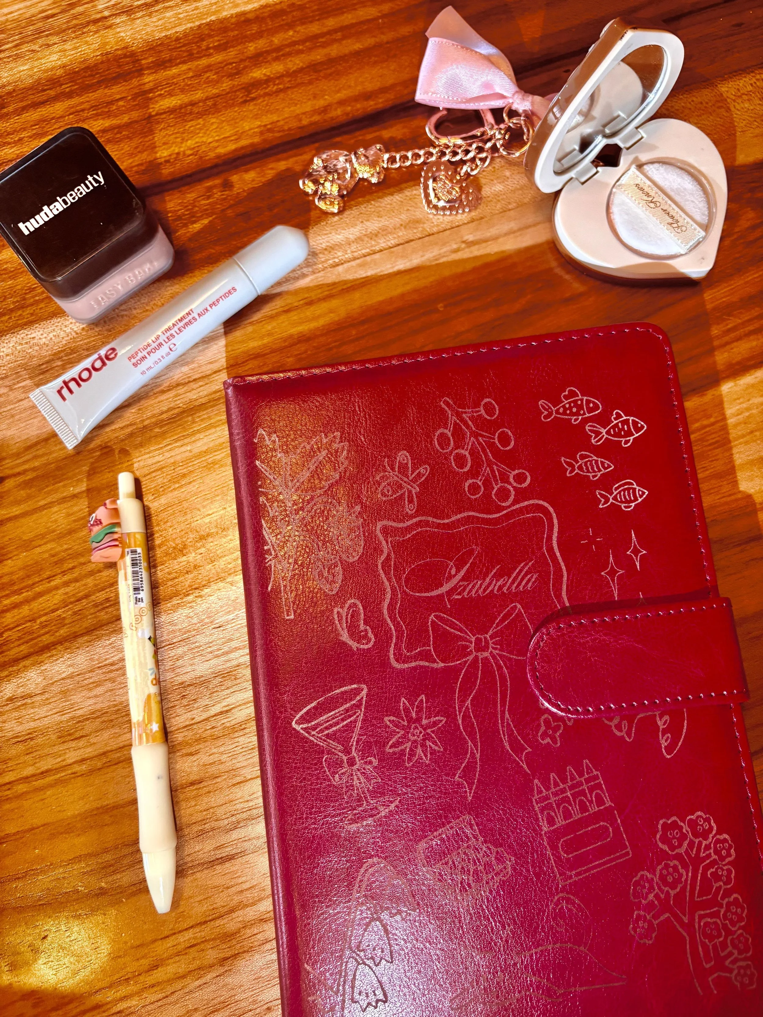 A wooden table with a red planner, a white pen, a small container of pink blush, a lip balm, a pink ribbon keychain, and an open pink mirror compact. The pink planner has white sketches of flowers, butterflies, a cake, and a handbag on the cover.